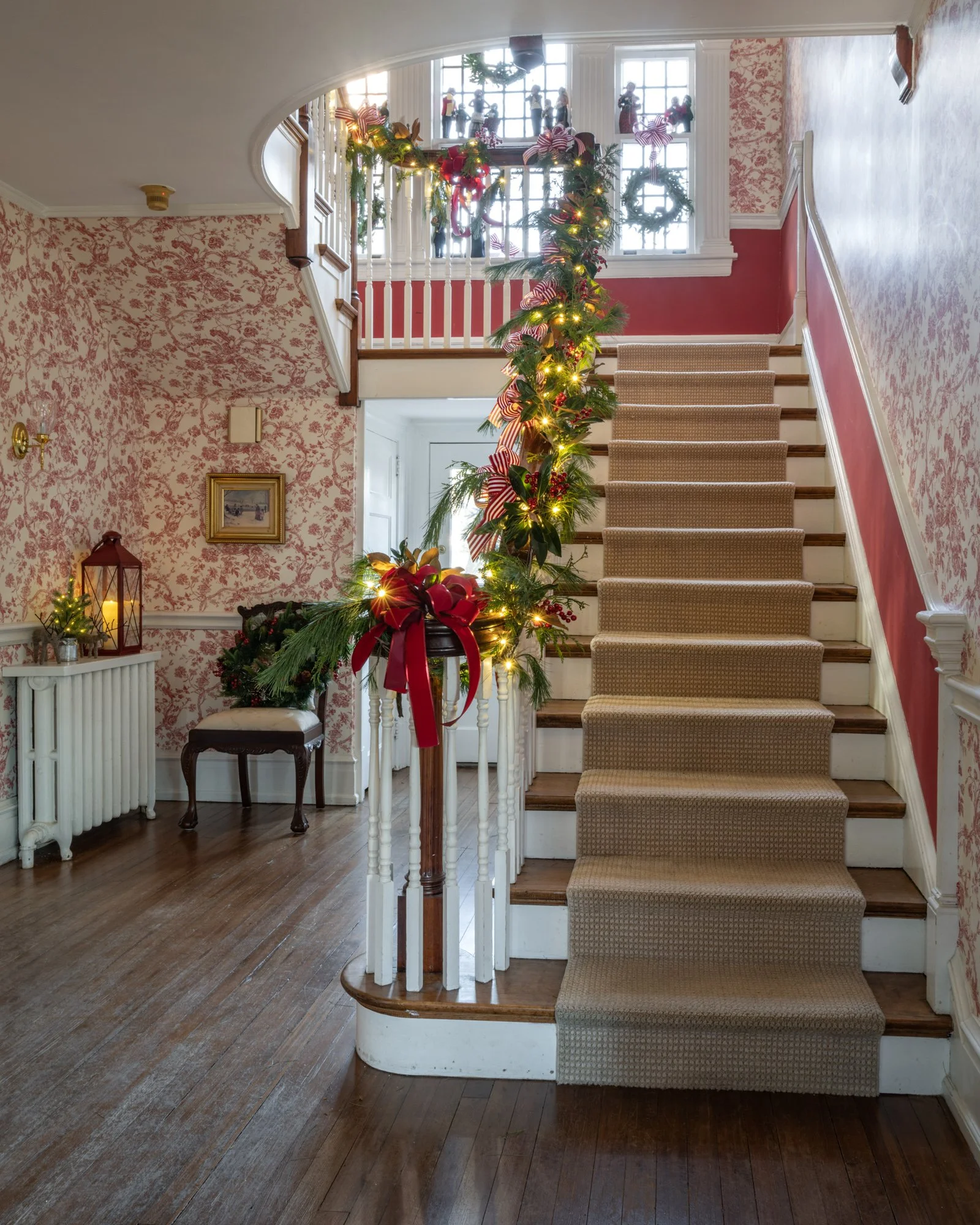 Interior view of a staircase decorated for Christmas with greenery, red bows, and lights. The staircase has wooden steps with beige carpet runners. In the background, a large window above the landing allows natural light into the space, with a railin