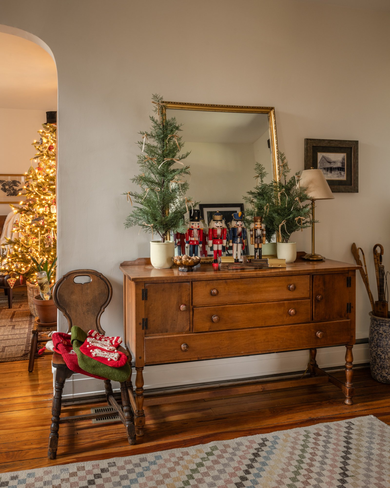 A wooden sideboard with three drawers and two cabinets, topped with two potted evergreen trees adorned with ribbons, nutcracker figurines, and a framed mirror. A chair with a Christmas stocking and a green hat is in front of the sideboard. In the bac