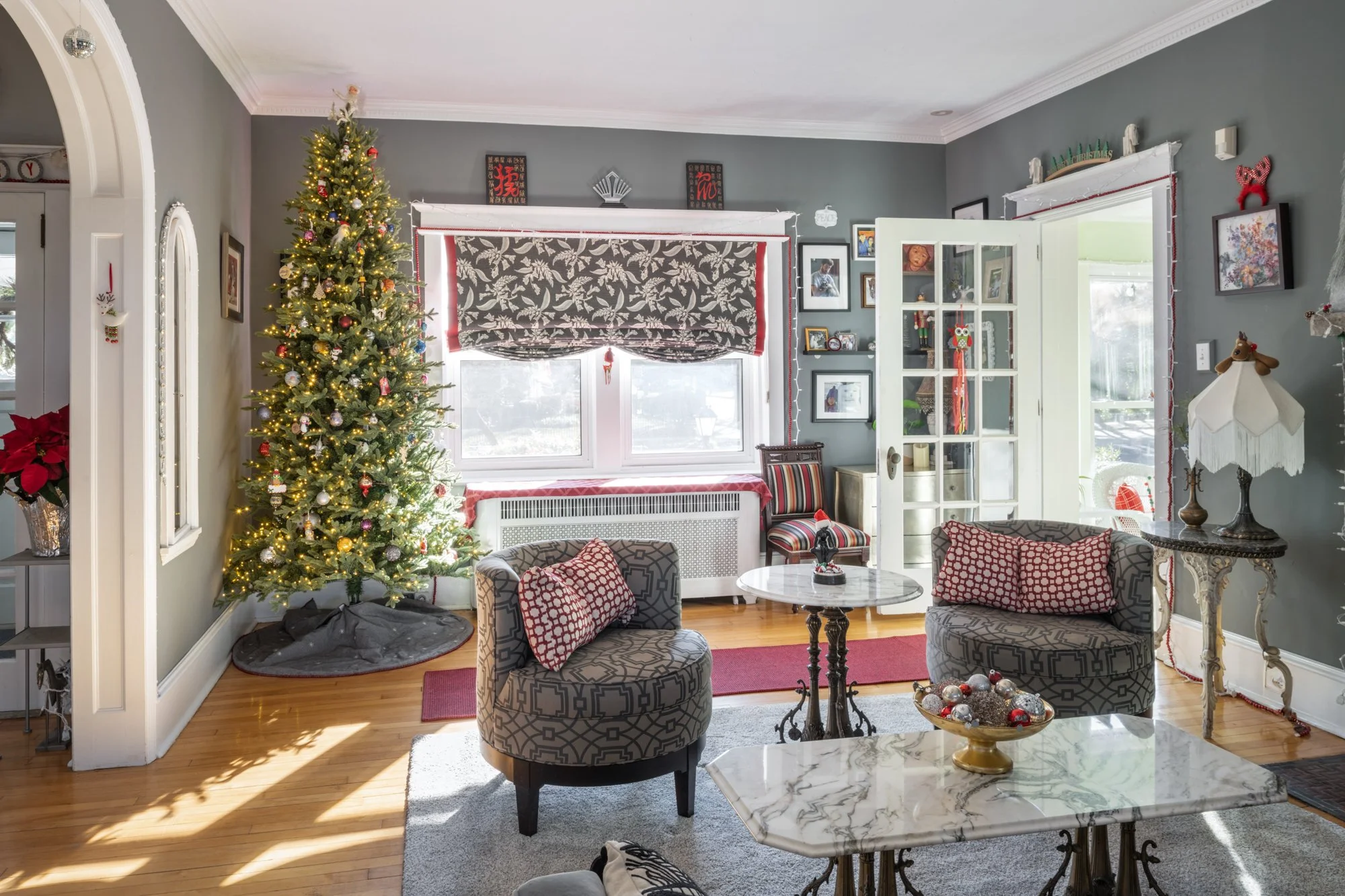 A living room decorated for Christmas with a tall, lit Christmas tree adorned with ornaments. The room has gray walls, crown molding, and a window with a dark patterned curtain. There are framed photos and artwork on the walls, a glass cabinet with h