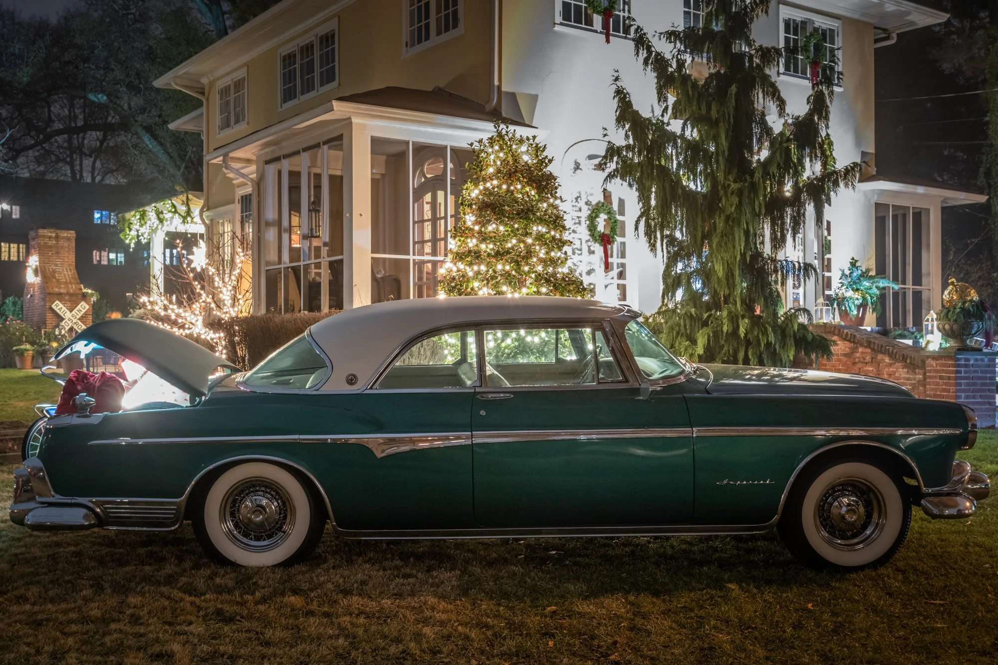 A classic green Cadillac convertible with white wall tires parked in front of a white house decorated for Christmas. The house has a Christmas tree with lights in the front yard, and wreaths with red bows hang on the house. There are string lights on