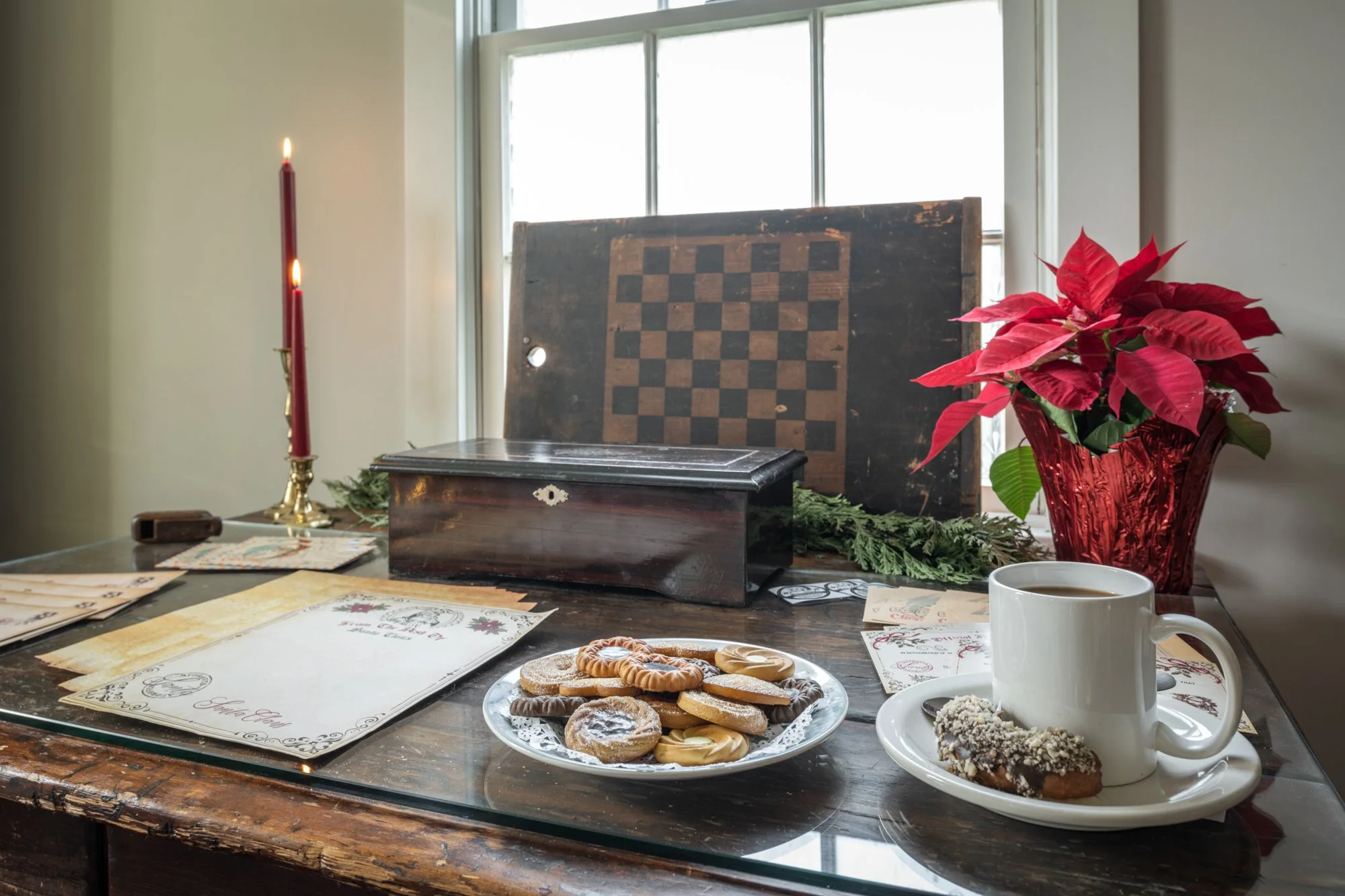 A dining table decorated for Christmas with a large plate of assorted cookies and a white mug of coffee, red poinsettia plant in a glass vase, lit red candles, chessboard, holiday cards, and a window in the background.