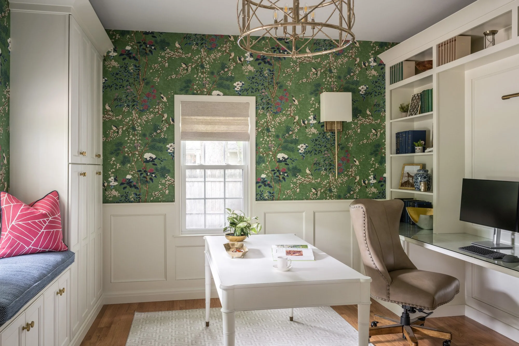 A cozy living room with a floral wallpapered accent wall, white built-in cabinets and shelves, a white coffee table with magazines, a beige armchair, a gray cushioned window seat with a pink and white patterned pillow, a tall white lamp, and a small 