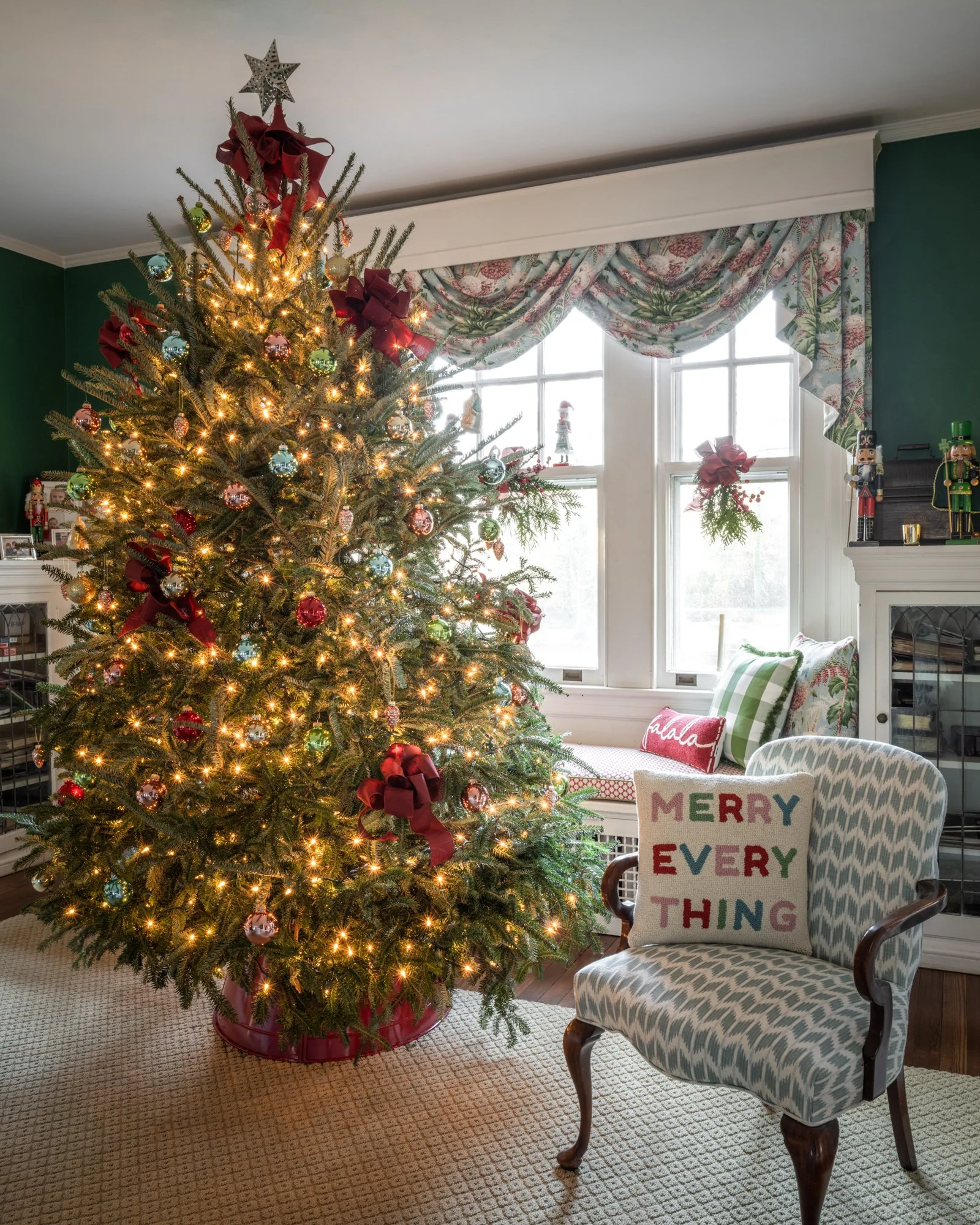 A decorated Christmas tree with lights, ornaments, ribbons, and a star on top, in a living room with sunlight coming through windows, and a patterned armchair with a pillow that says 'MERRY EVERY THING' next to it, along with holiday decor and nutcra