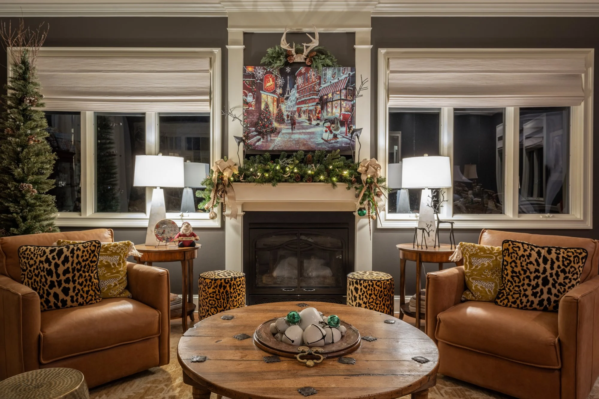 Living room decorated for Christmas with two leather sofas, a round wooden coffee table with ornaments, a fireplace mantel with greenery and bows, a picture of a Christmas street scene above the mantel, two lamps, and large windows with white shades.