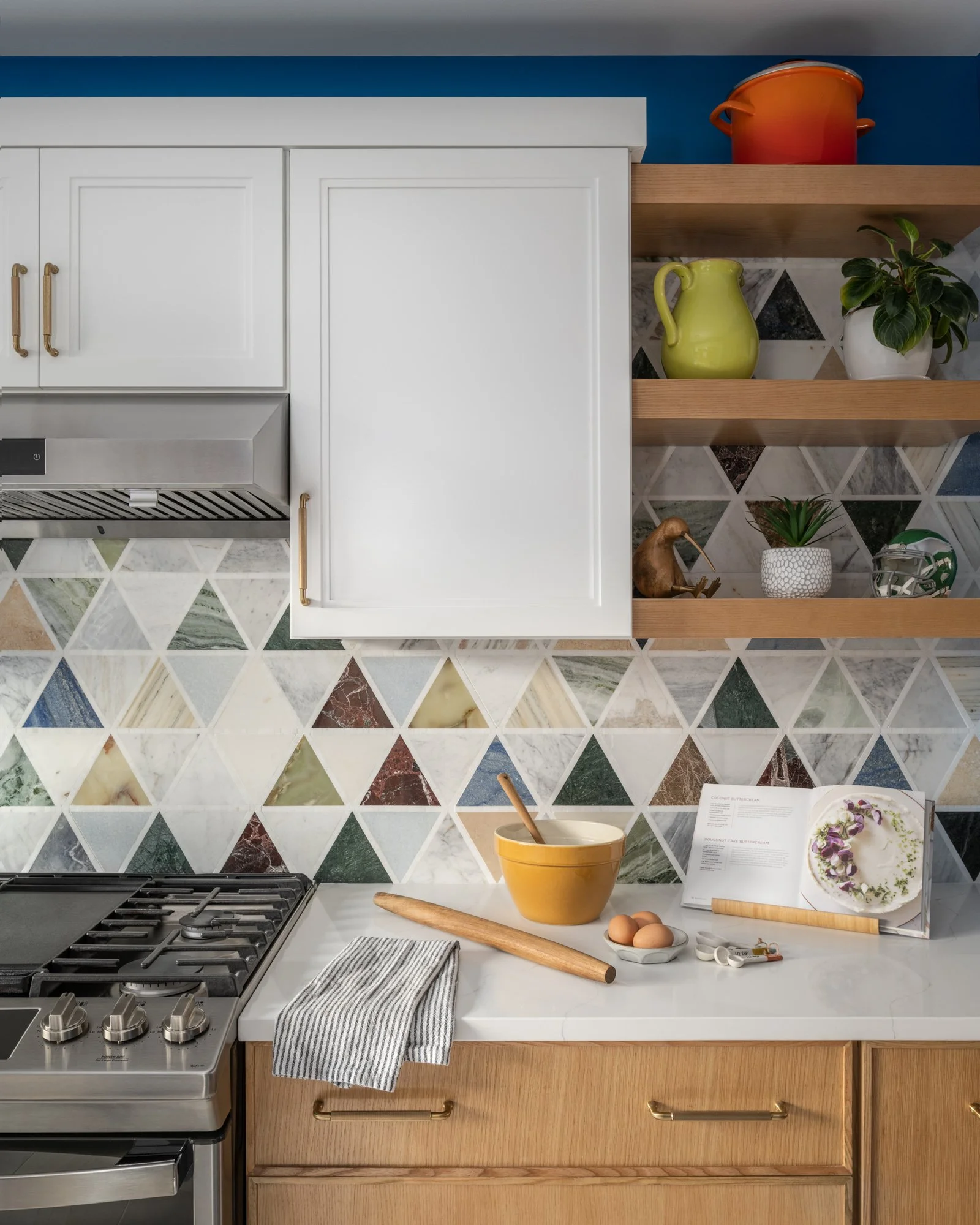 Kitchen with a gas stove, wooden cabinets, and open shelves displaying a green pitcher, potted plant, red pot, cookbook, and decorative items. The countertop has eggs, a yellow mixing bowl, rolling pin, kitchen utensils, and a dishcloth in front of a