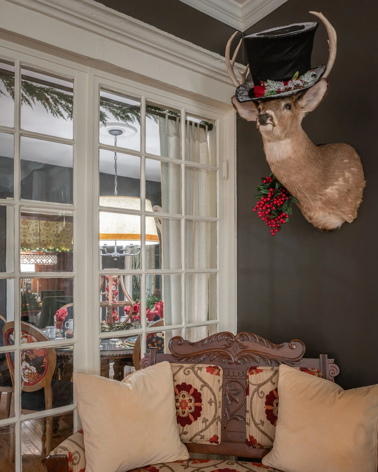 Living room with a decorative deer head mounted on the wall, wearing a black top hat with Christmas decorations and antlers. There is a window with white frames, and a dining area with table settings and holiday decorations visible through the window