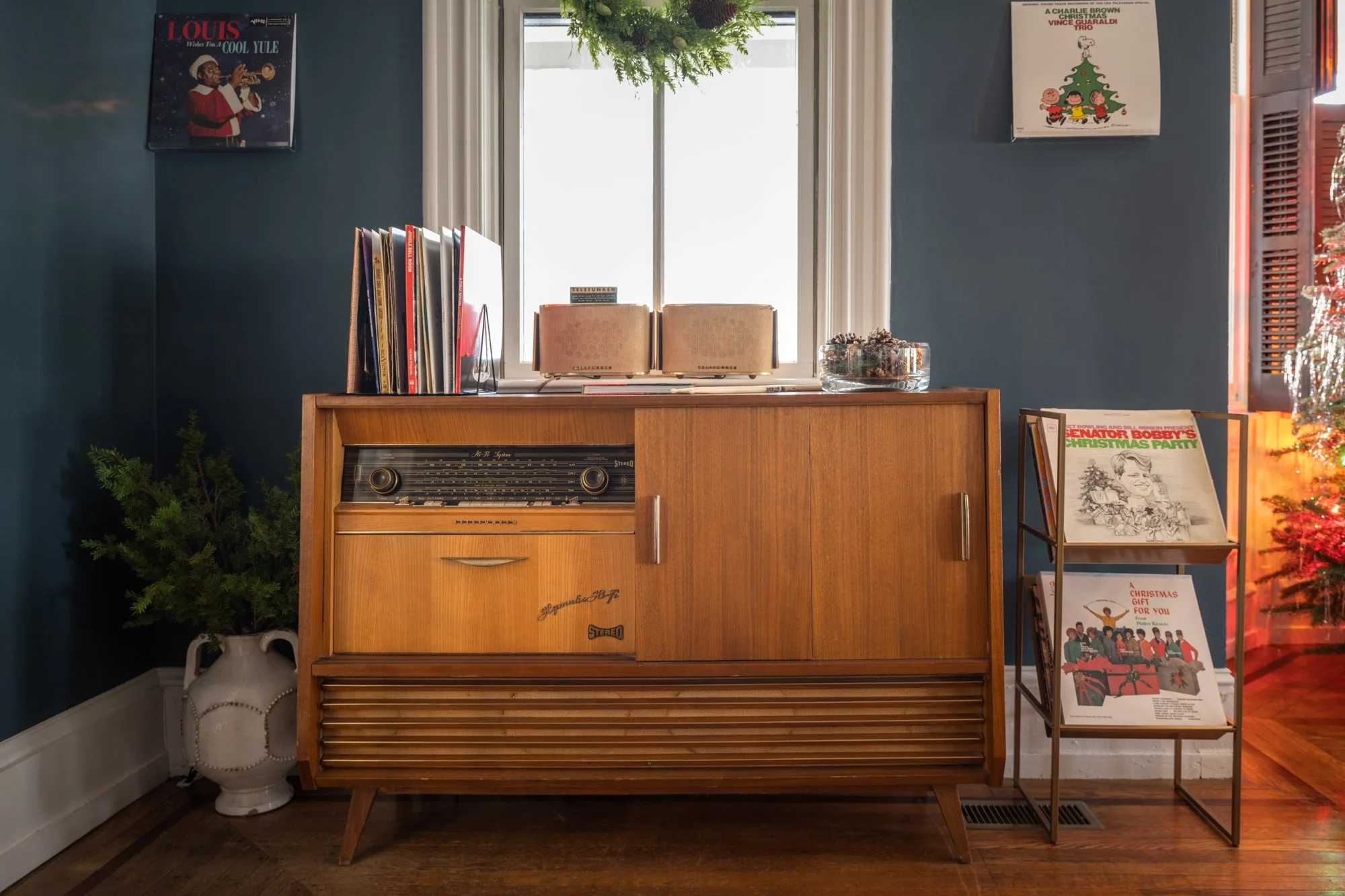 A wooden vintage stereo cabinet with books and magazines on top against a blue wall, with framed album covers and a window with greenery outside. A small potted plant is on the left, and a metal magazine rack with more magazines is on the right side.
