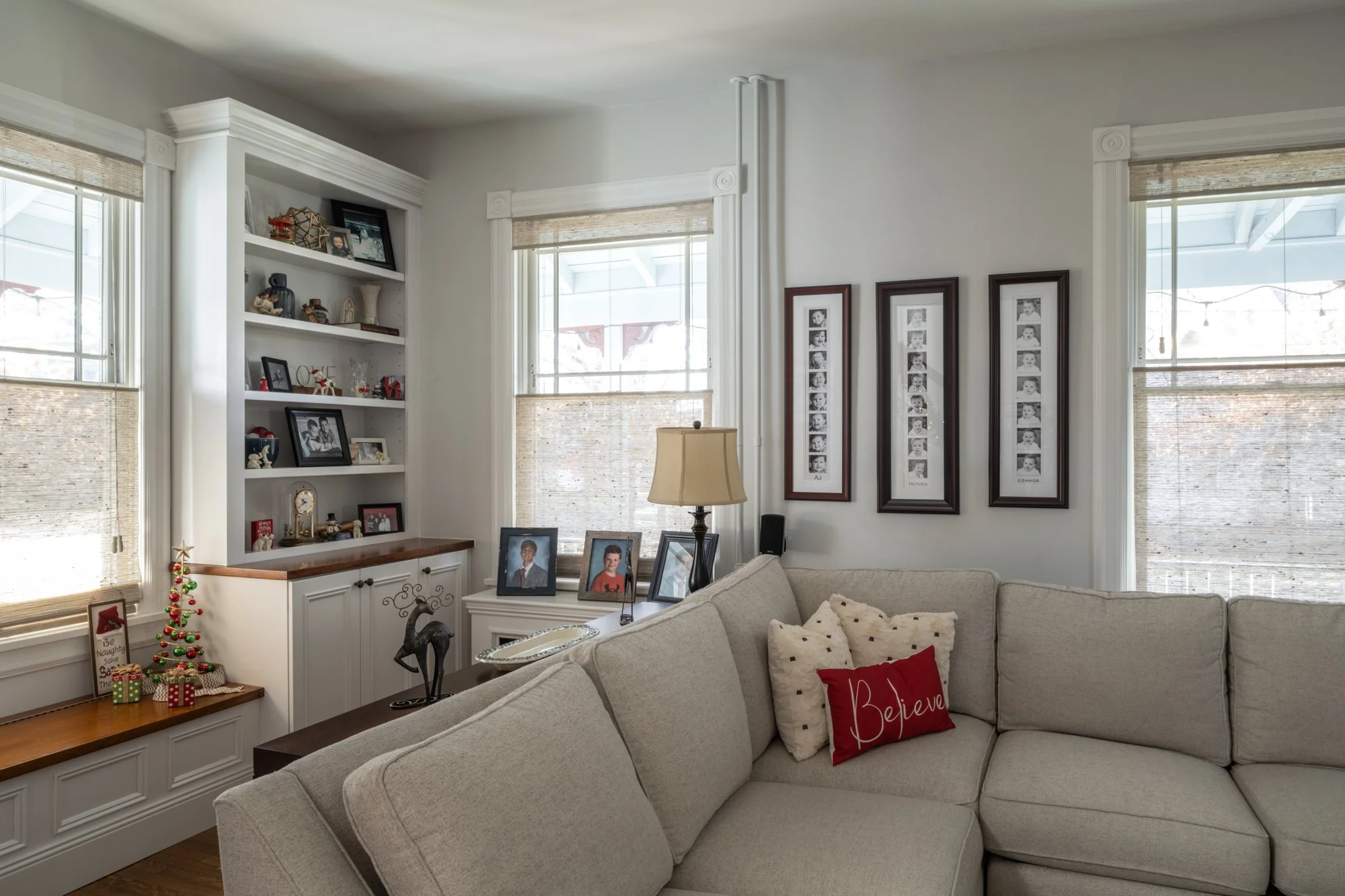 A living room with white walls, a beige sectional sofa with pillows, and a white built-in bookshelf with framed photos and decorative items. There are windows with woven shades, and Christmas decorations including a small tree and presents on a nearb