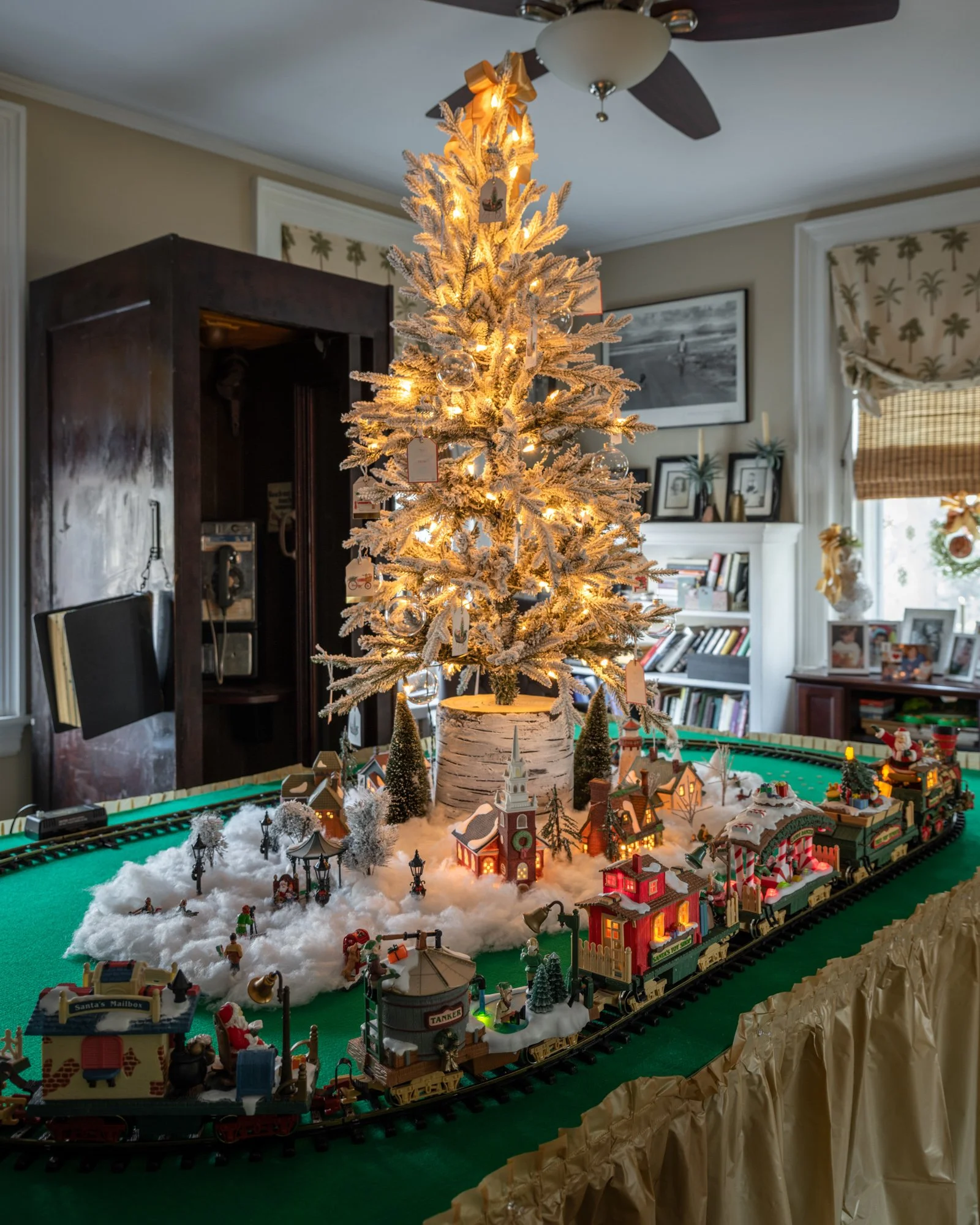 A decorated Christmas tree with lights inside a living room. A snow-themed miniature train set surrounding the tree displays tiny buildings, trees, and figurines. The room has bookshelves, framed pictures, and a ceiling fan.