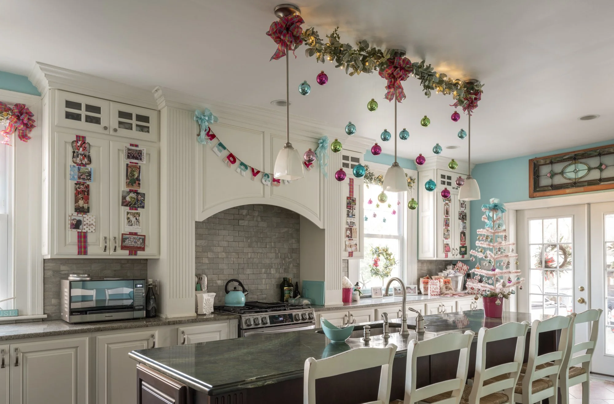 Kitchen decorated for Christmas with ornaments hanging from the ceiling, a small Christmas tree, and holiday banners and photos on the walls.