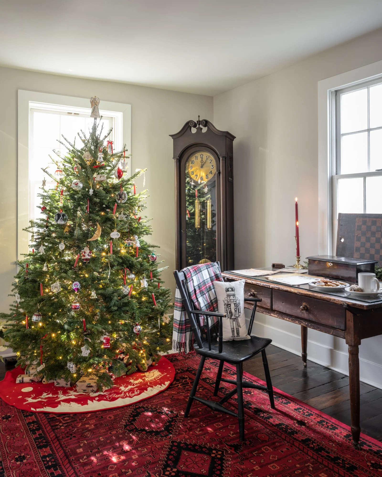 A decorated Christmas tree with ornaments and lights, a grandfather clock, a table with a red candle, and a chair with a holiday pillow, all in a room with white walls, large windows, and a red rug.