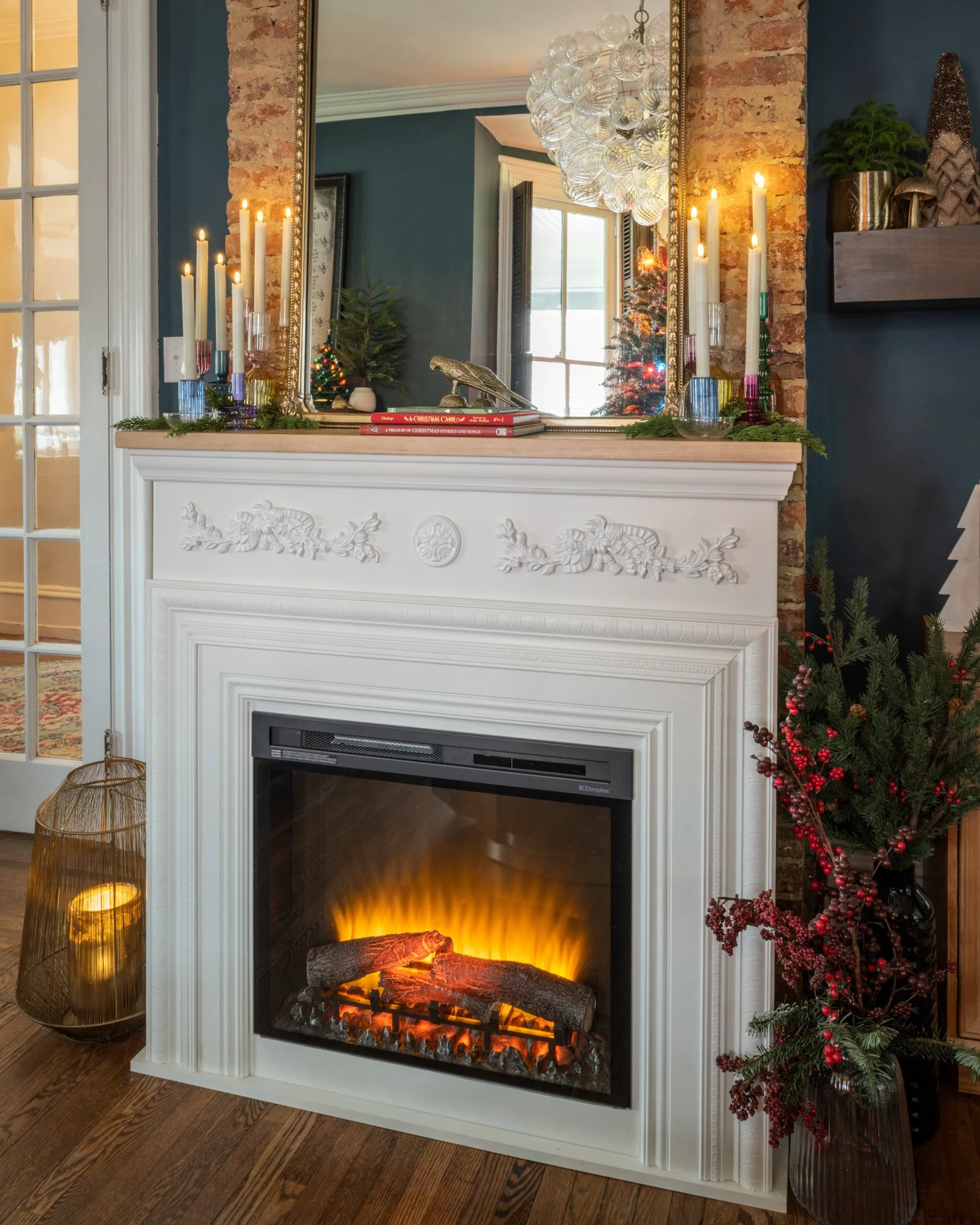 A decorated fireplace mantel with lit candles, a small pine tree, holiday books, and a gold bird figurine reflected in a large mirror. To the right of the fireplace, there is a red berry-filled plant, and to the left, a gold wire lantern with a candl