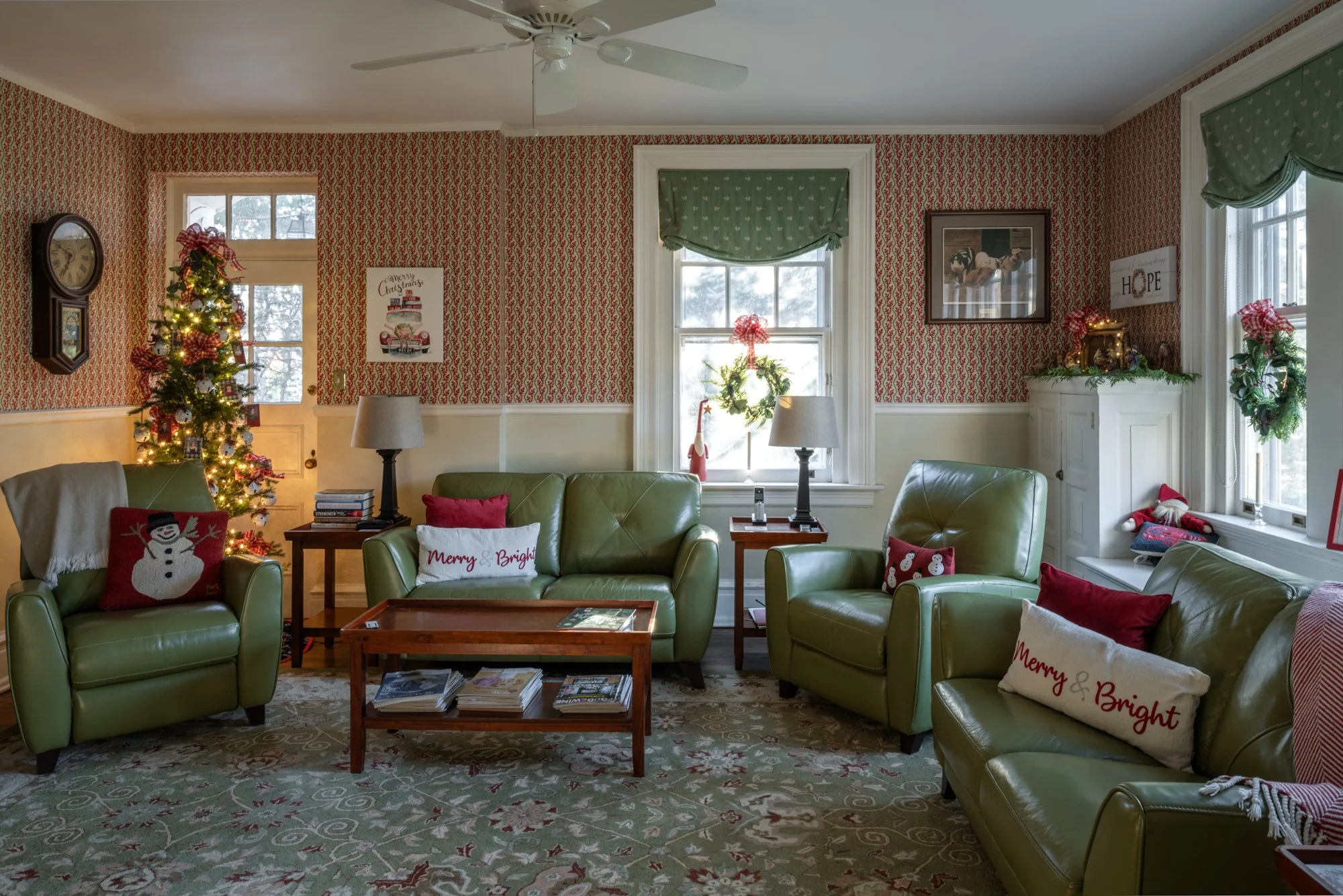 Living room decorated for Christmas with a lit Christmas tree, stockings on a white fireplace, holiday pillows, and framed artwork on patterned wallpaper.