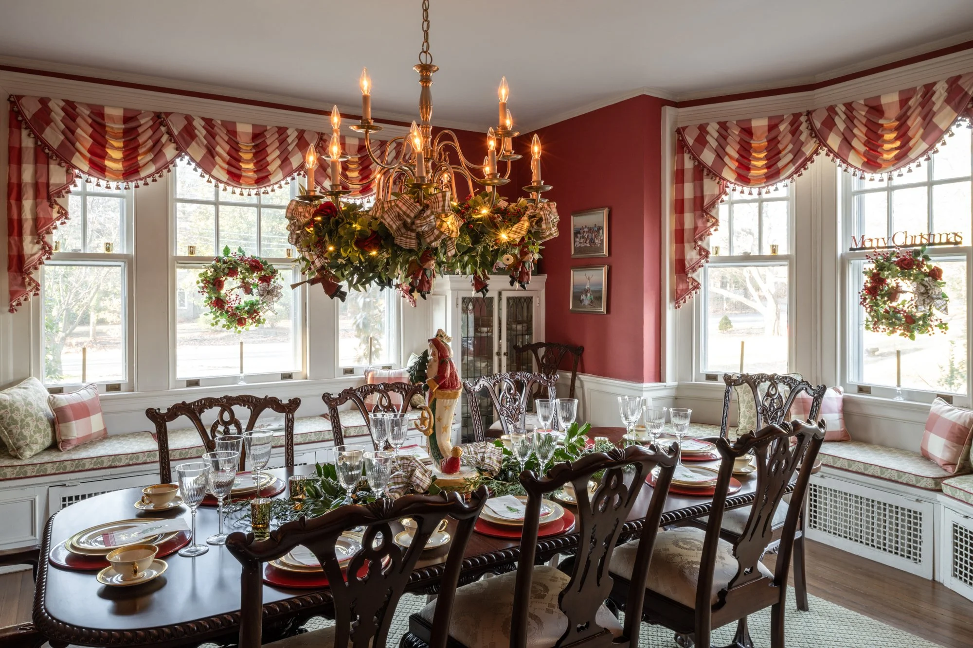 A dining room decorated for Christmas with a chandelier, holiday wreaths, and window curtains with red and white patterns.