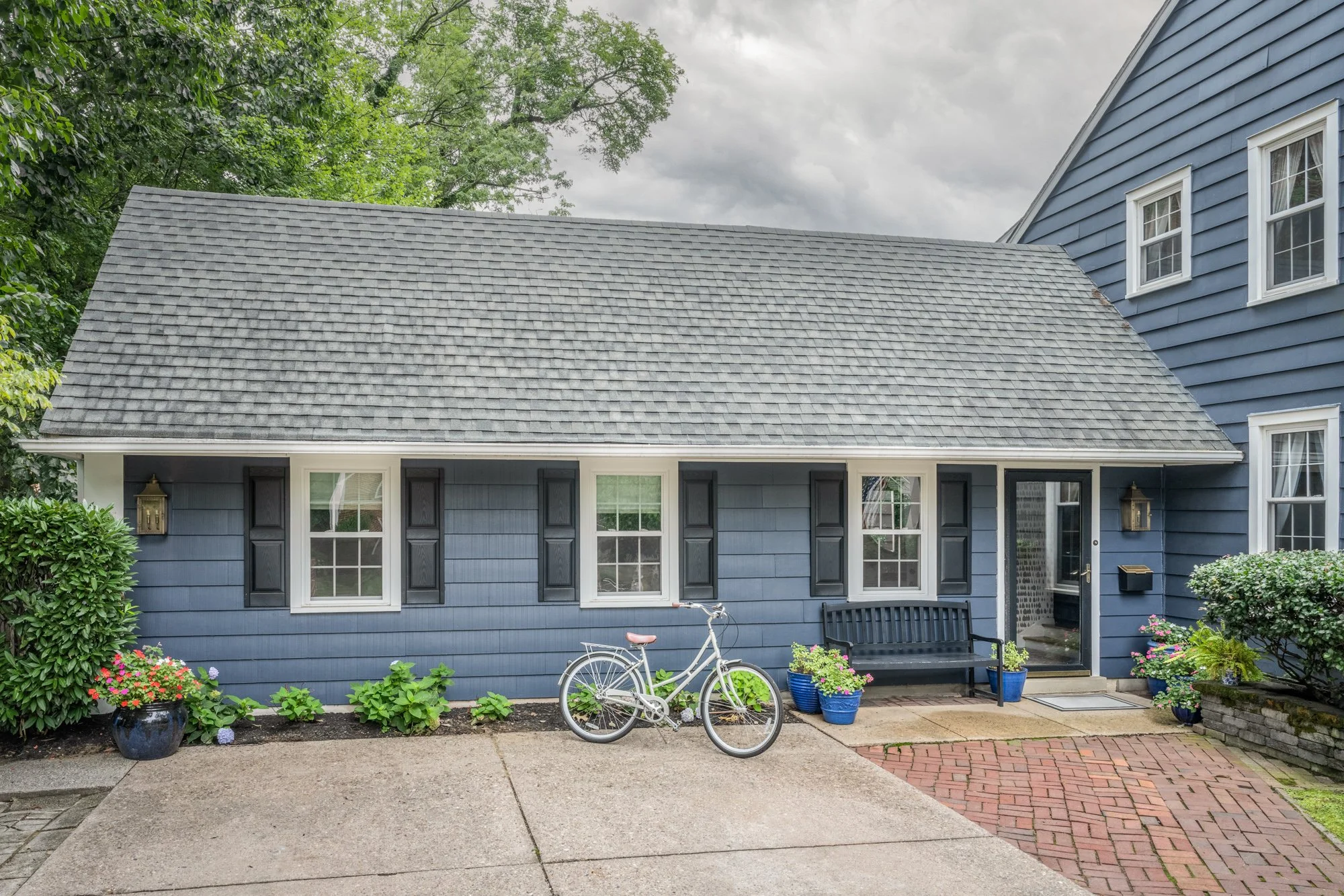 Front of a blue house with a sloped gray shingle roof, white-framed windows, a black door, and a black bench outside. There are potted plants and a white bicycle in the garden area in front. The sky is overcast and there are trees in the background.