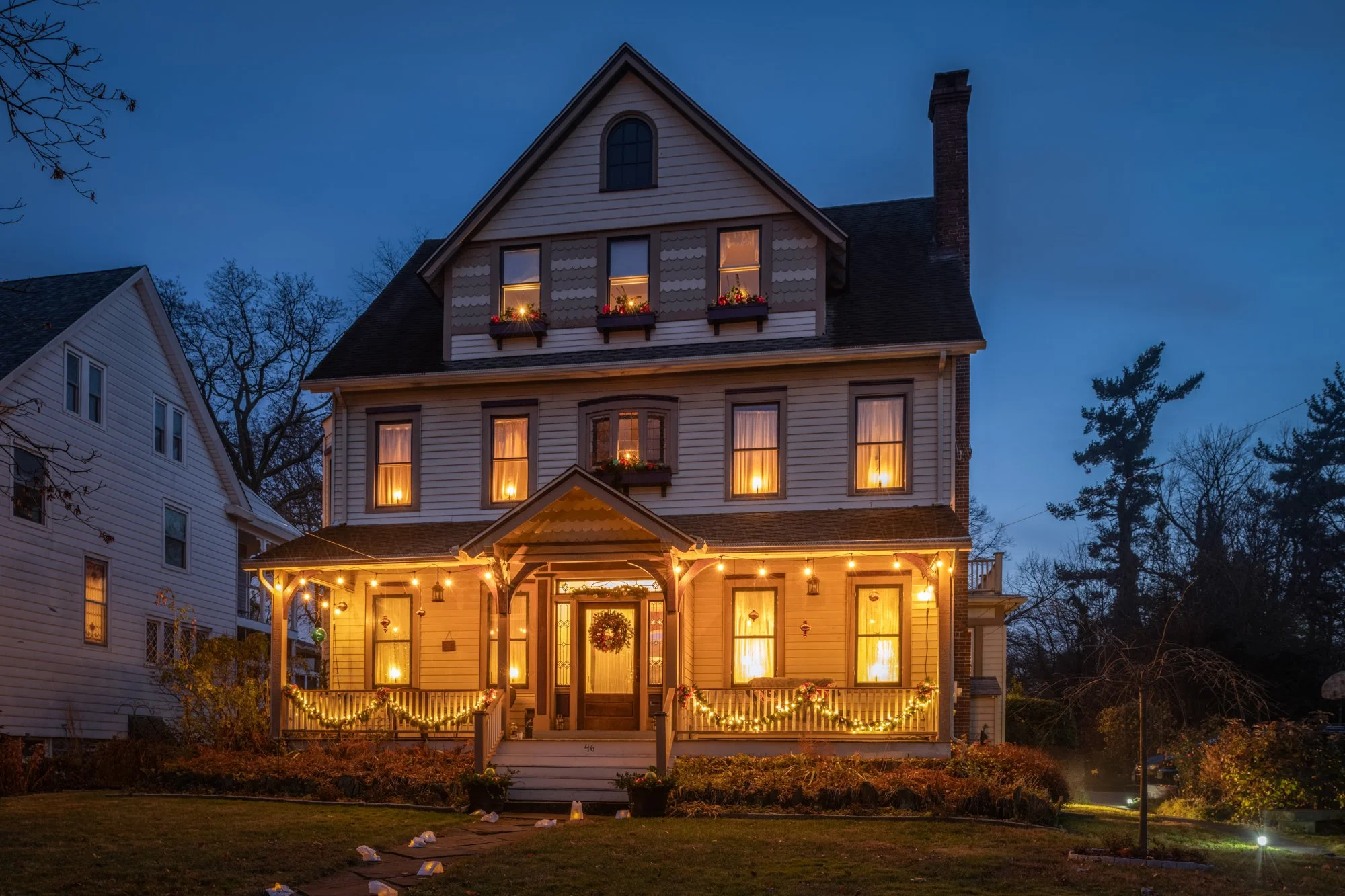 A large, multi-story house decorated with Christmas lights and wreaths, with warm yellow lights glowing from the windows, during dusk or early evening.