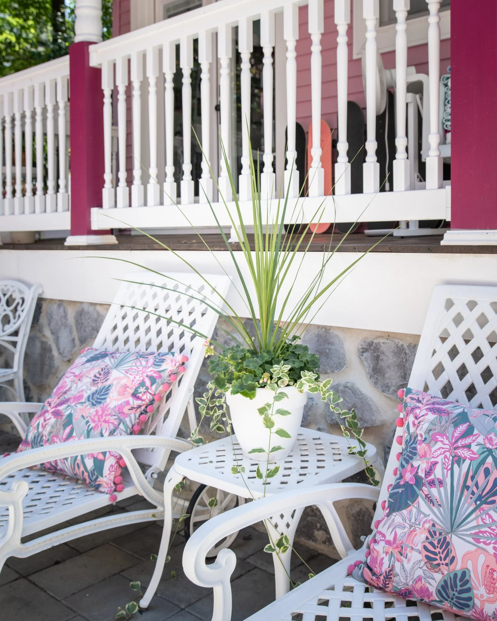 A white round metal table with a large potted green plant in the center, surrounded by white metal chairs with pink and purple floral cushions, on a stone patio with a pink house and white railing in the background.