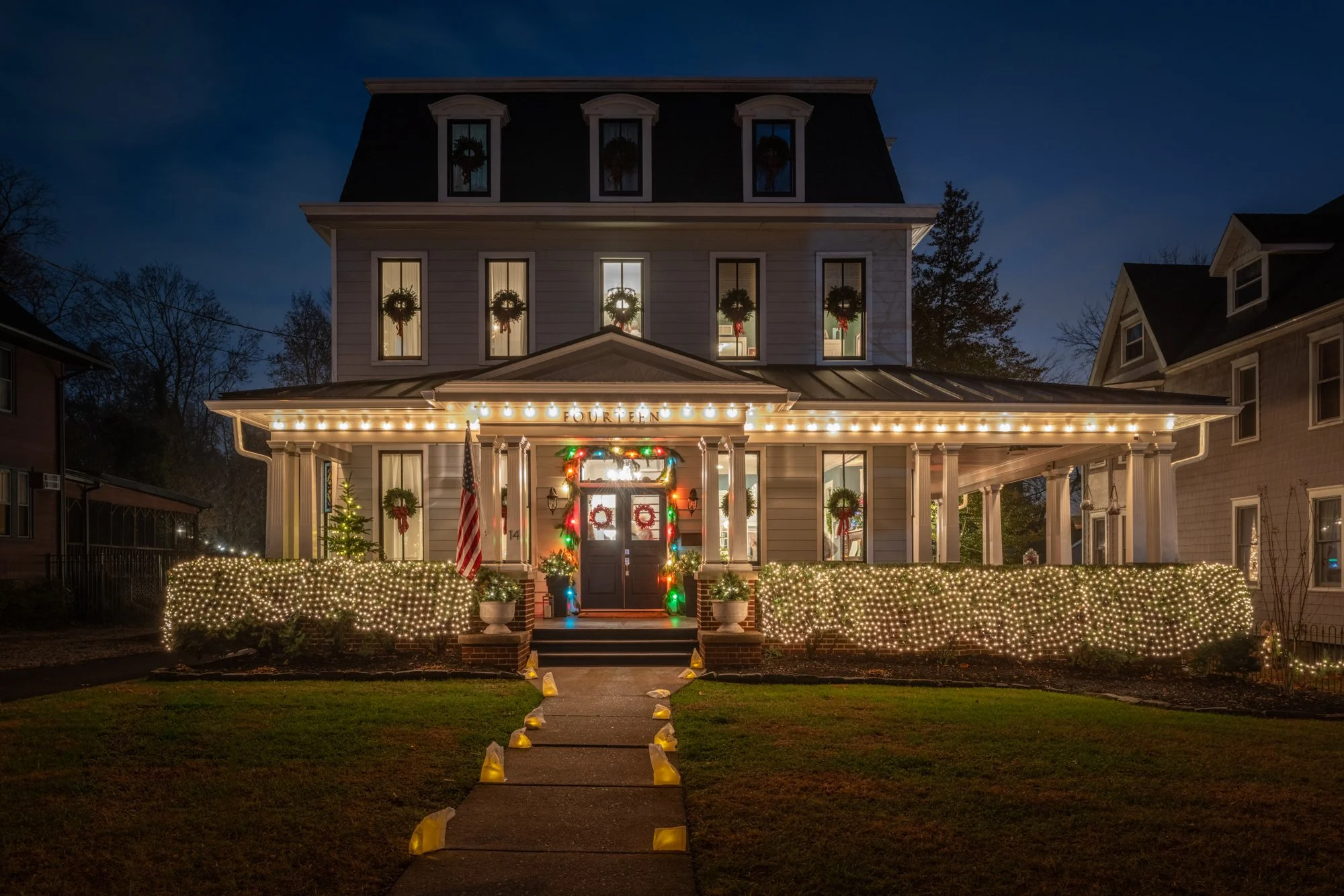 A large house decorated with Christmas lights and wreaths, with a pathway illuminated by small lights leading to the front door, which is decorated with colorful lights and ornaments. The house has multiple stories, with wreaths on the windows, a ban