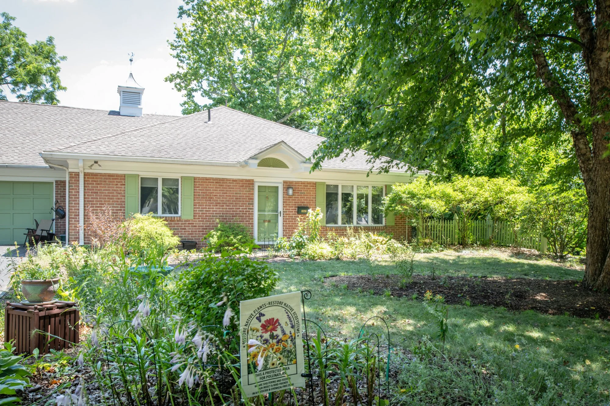 A house with a brick exterior, green shutters, and a gray roof surrounded by trees and a well-maintained garden with flowering plants. There is a sign in the garden about native plant restoration and pollinator protection.