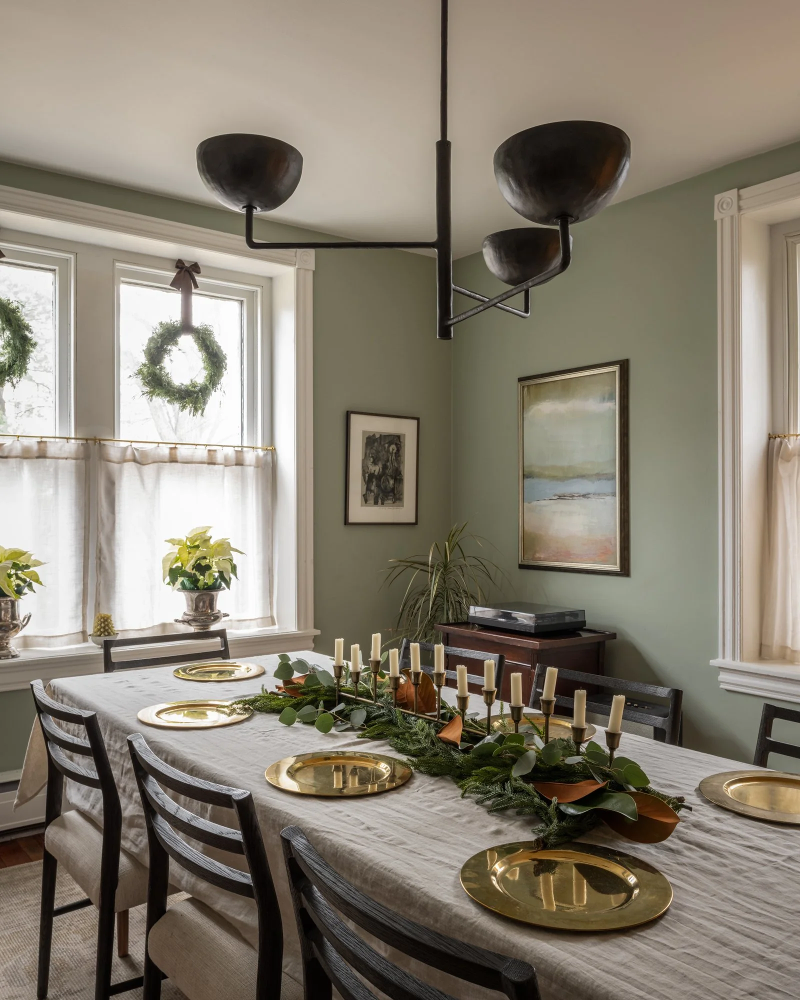 A dining room with a table set for a holiday meal, decorated with greenery and candles, with large windows and wall art.