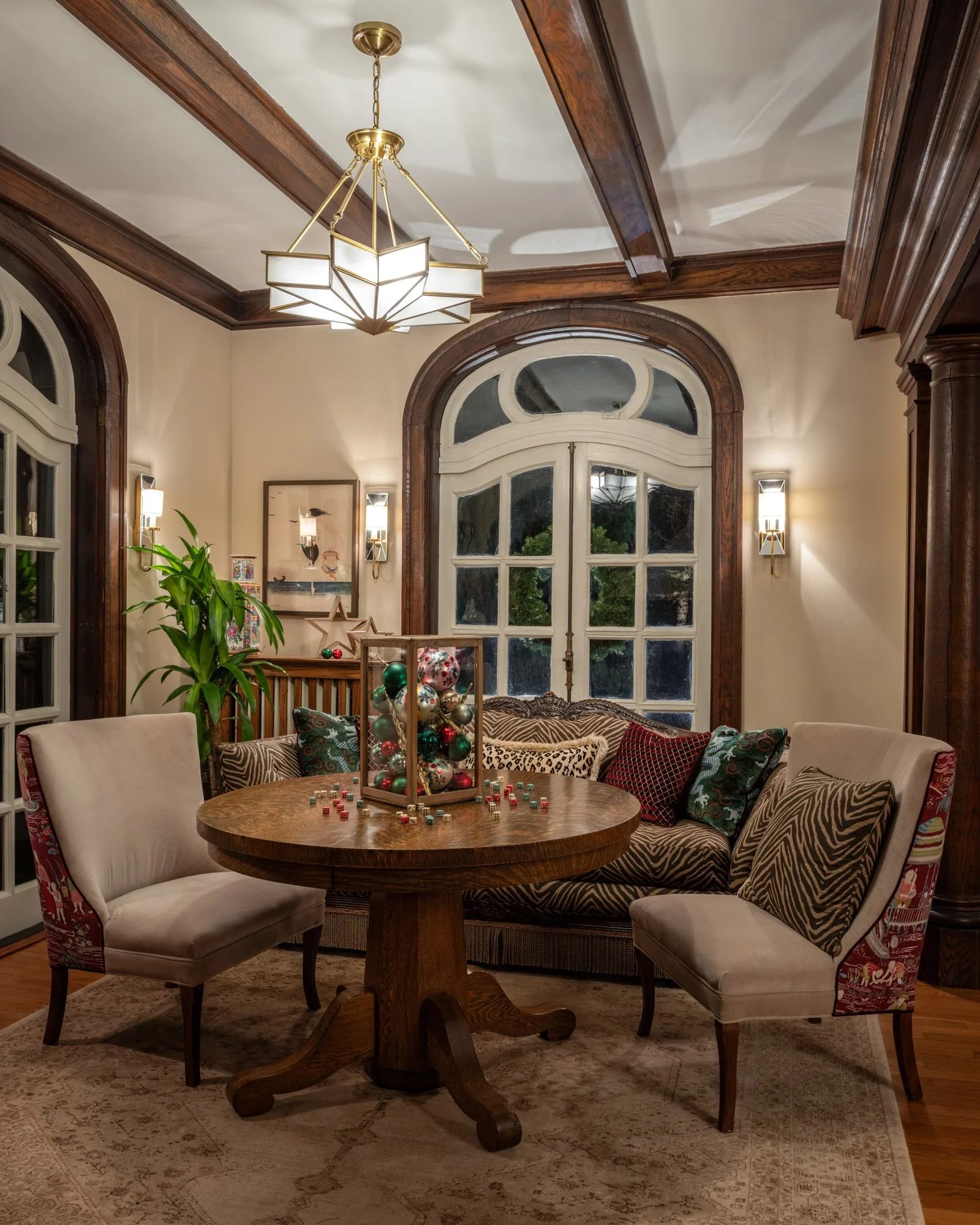 A cozy living room with a wooden round table surrounded by colorful and neutral armchairs and sofa, a Christmas decoration in a gold cage on the table, a large window, a chandelier, and wall-mounted lights.