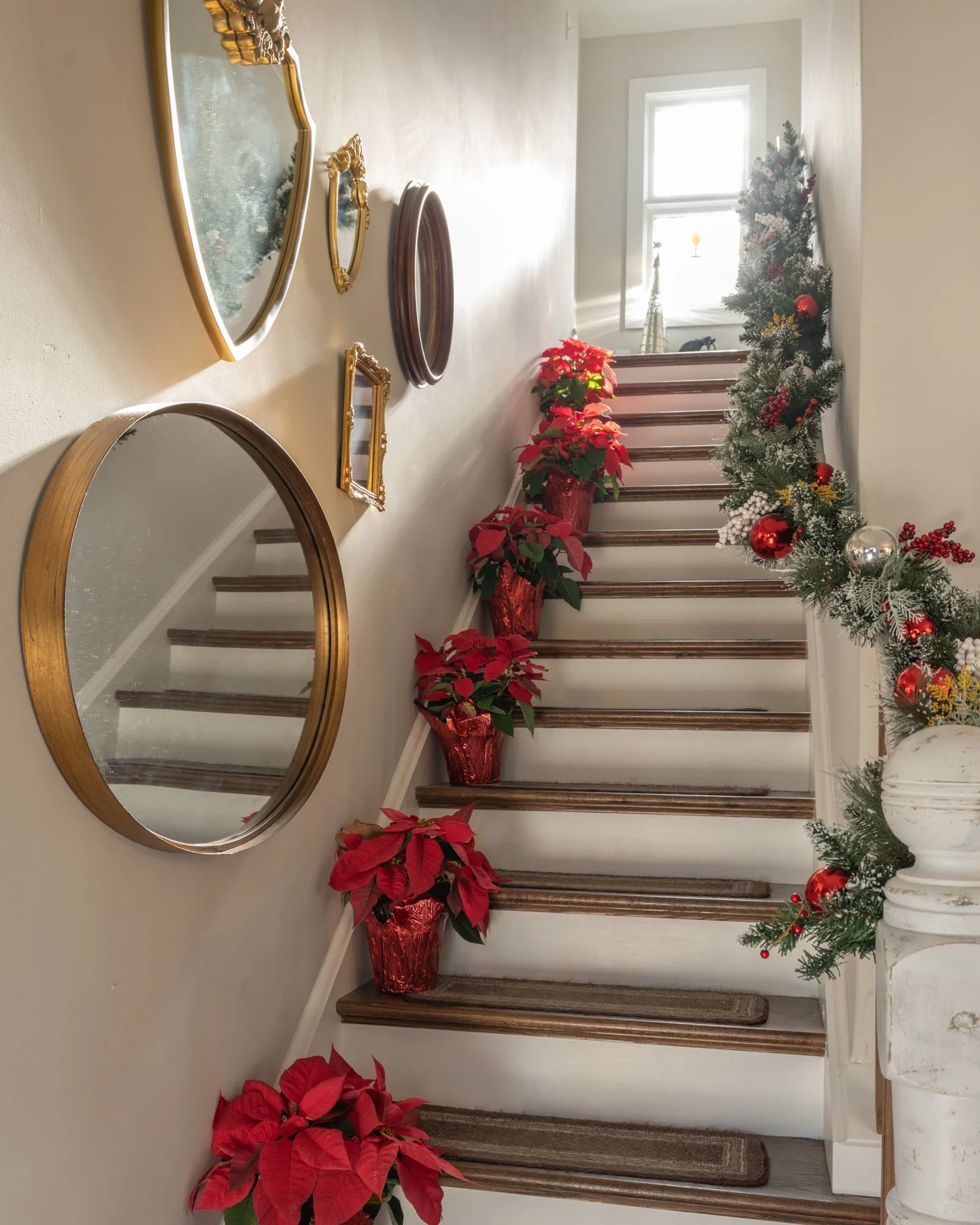 Decorated staircase with Christmas garland, red poinsettias, and ornaments, with framed mirrors on the wall and a window at the top.