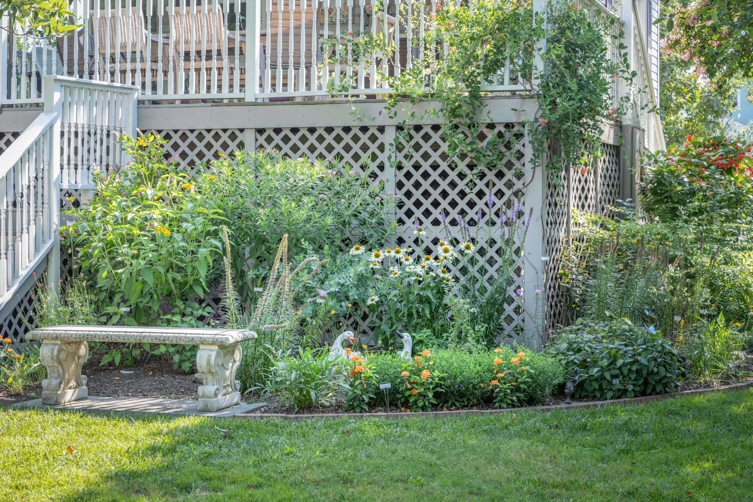 A lush garden with colorful flowers and green foliage in front of a white fence and staircase leading to a balcony, with decorative garden sculptures of ducks near the plants.