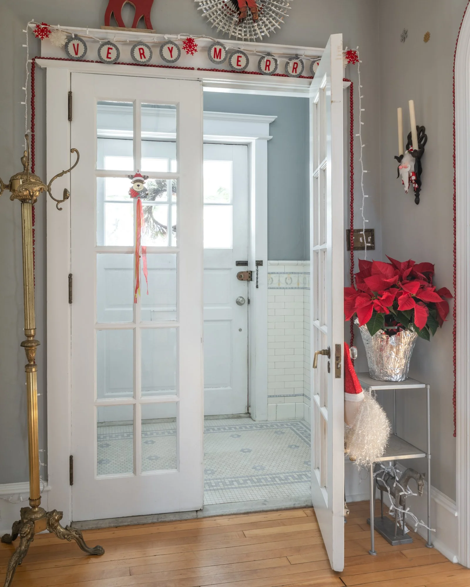 A decorated entrance of a home with Christmas and holiday decorations, including a "Very Merry" banner, poinsettia plant, Santa hat, and furry white decoration on a small table, with a glass-paneled door leading to a porch.
