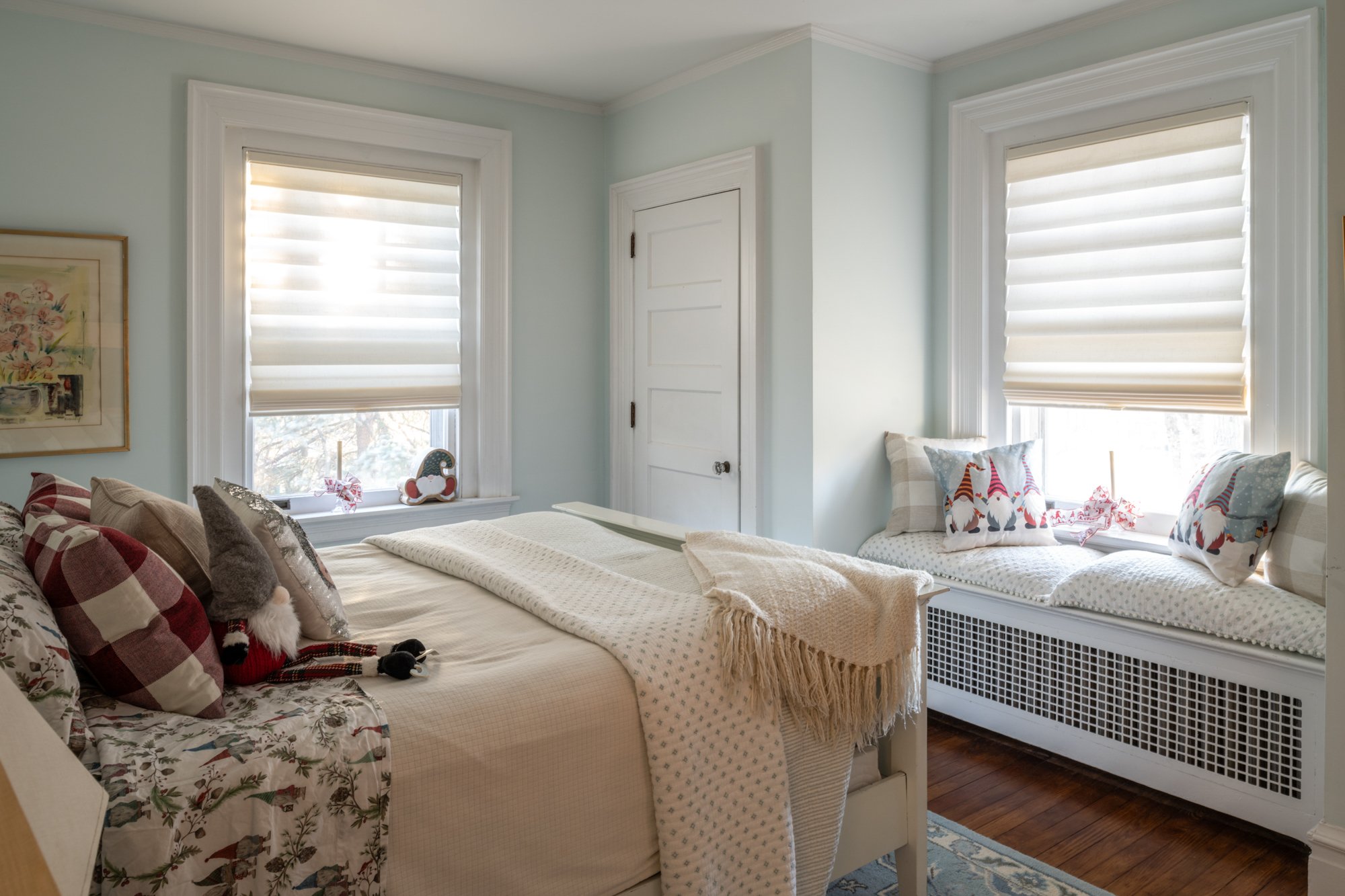 A cozy bedroom with two beds, decorated for Christmas, featuring holiday-themed pillows and plush toys, with bright windows and light blue walls.