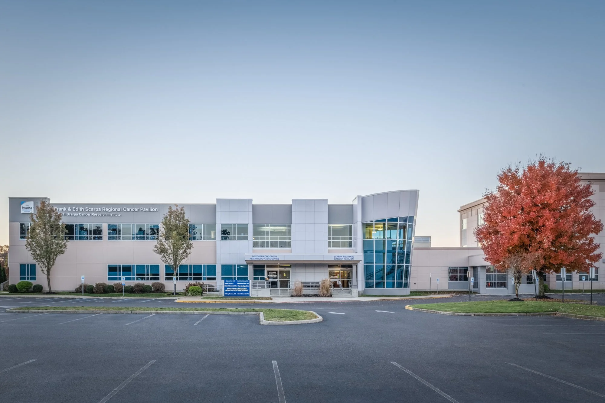 Exterior view of a modern medical building with some trees, parking lot, and a colorful autumn tree on the right. The signage indicates it is part of the Frank & Edith Scarpa Regional Cancer Pavilion and related cancer research and treatment faciliti