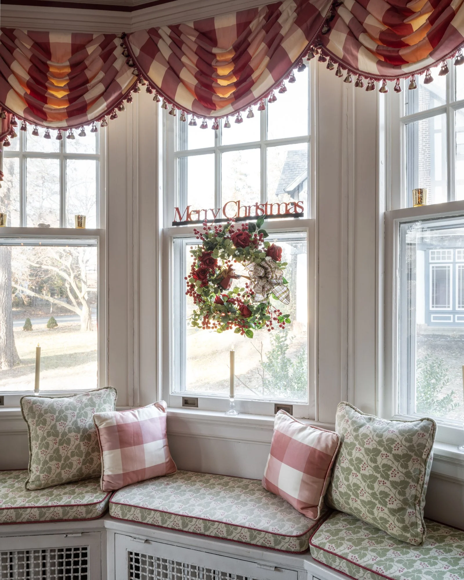 A cozy window seat with floral and checkered cushions, surrounded by large windows, decorated with a Christmas wreath, a 
