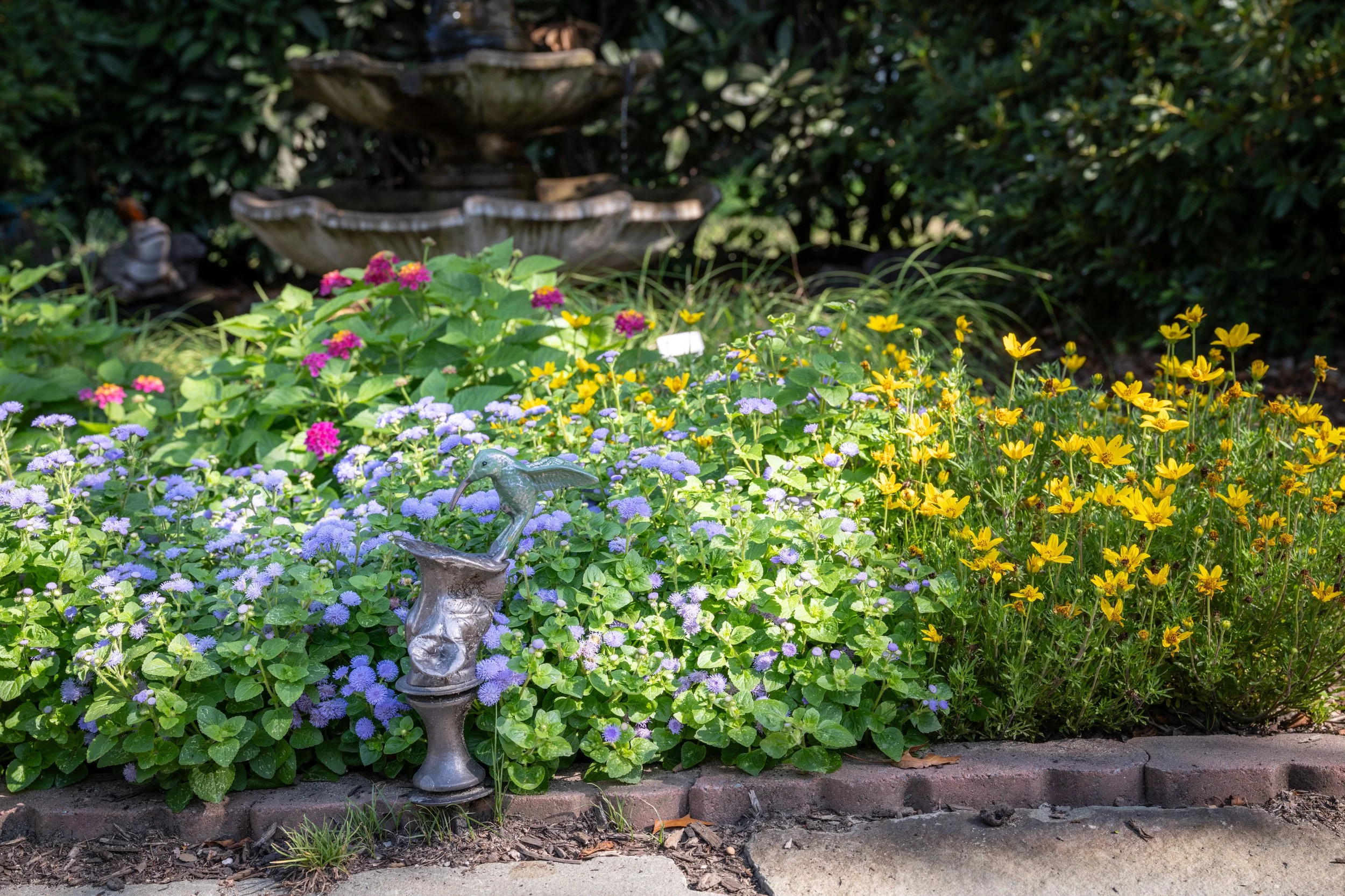 A garden with colorful flowers including purple, pink, yellow, and blue, a bird-shaped garden ornament, a stone pathway, and a two-tiered fountain in the background surrounded by green shrubs.