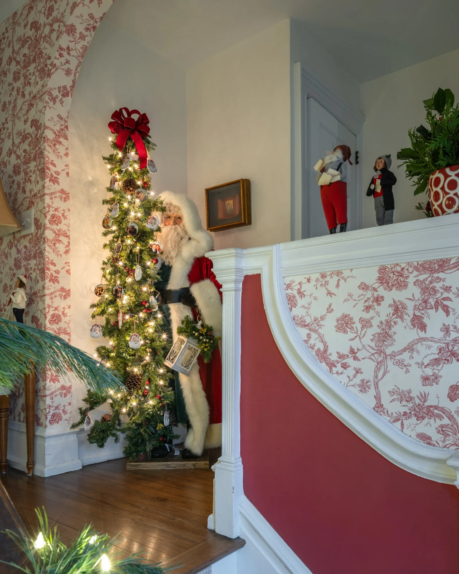 Decorated Christmas tree with ornaments and lights, Santa Claus figurine dressed in a red coat and hat, and holiday dolls, inside a house with red and white wallpaper and a wooden floor.