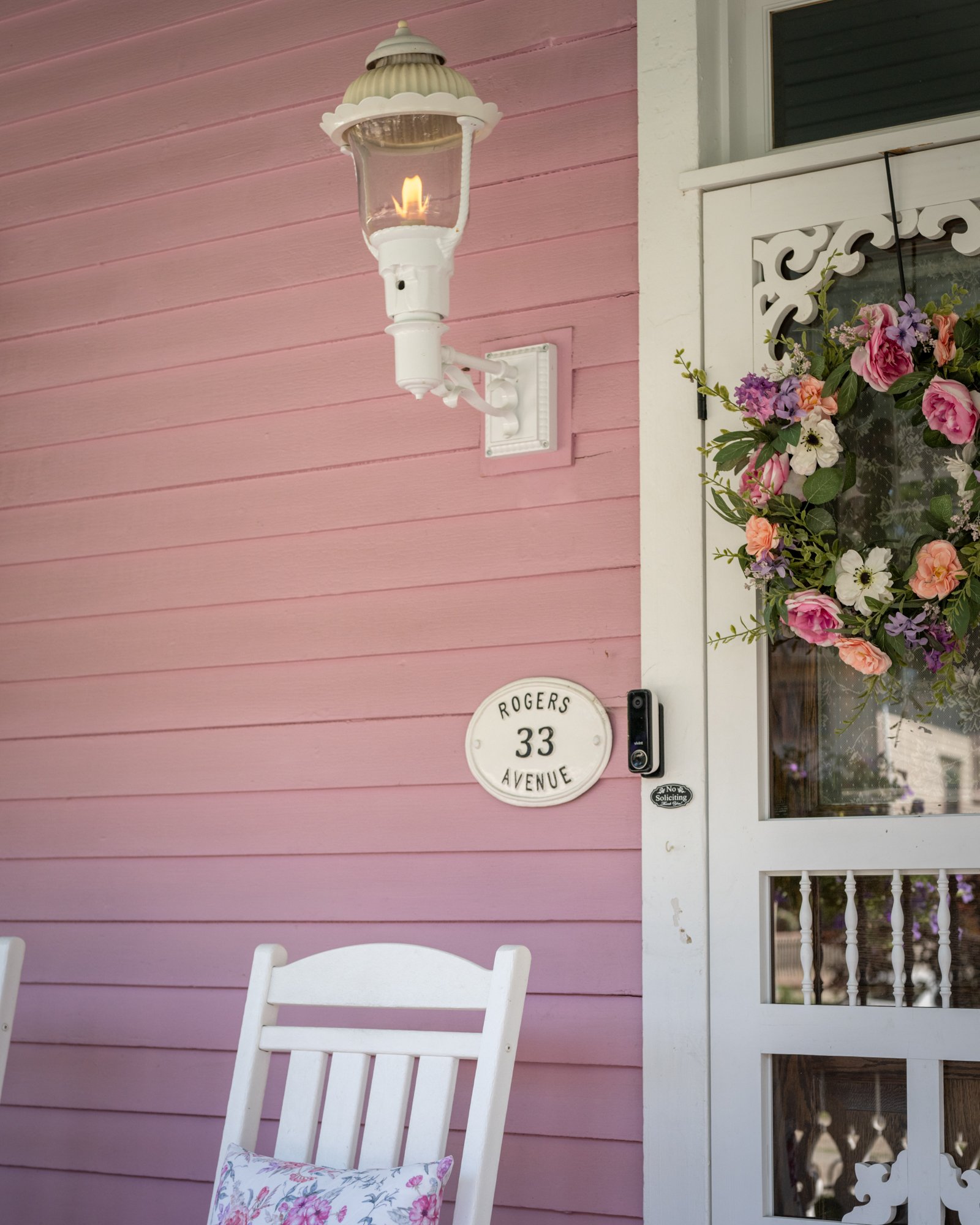 A white porch chair with a floral cushion in front of a pink wooden house, featuring a street sign that reads 'Rogers 33 Avenue', a white vintage outdoor lamp with a flickering flame, and a wreath of pink, purple, and white flowers on the door.