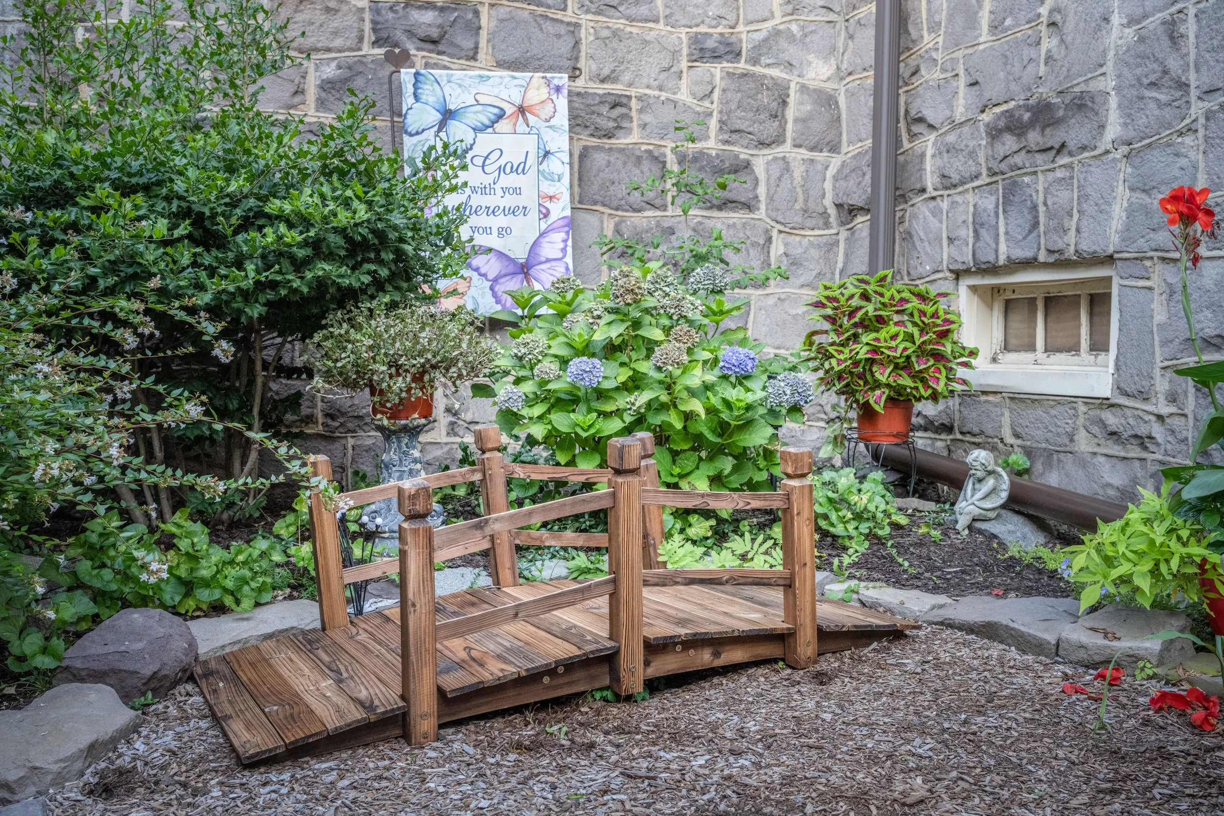 A small garden with a wooden bench, various flowering plants including hydrangeas and coleus, a garden statue, and a decorative sign with butterflies that reads 'God is with you wherever you go,' against a stone wall.