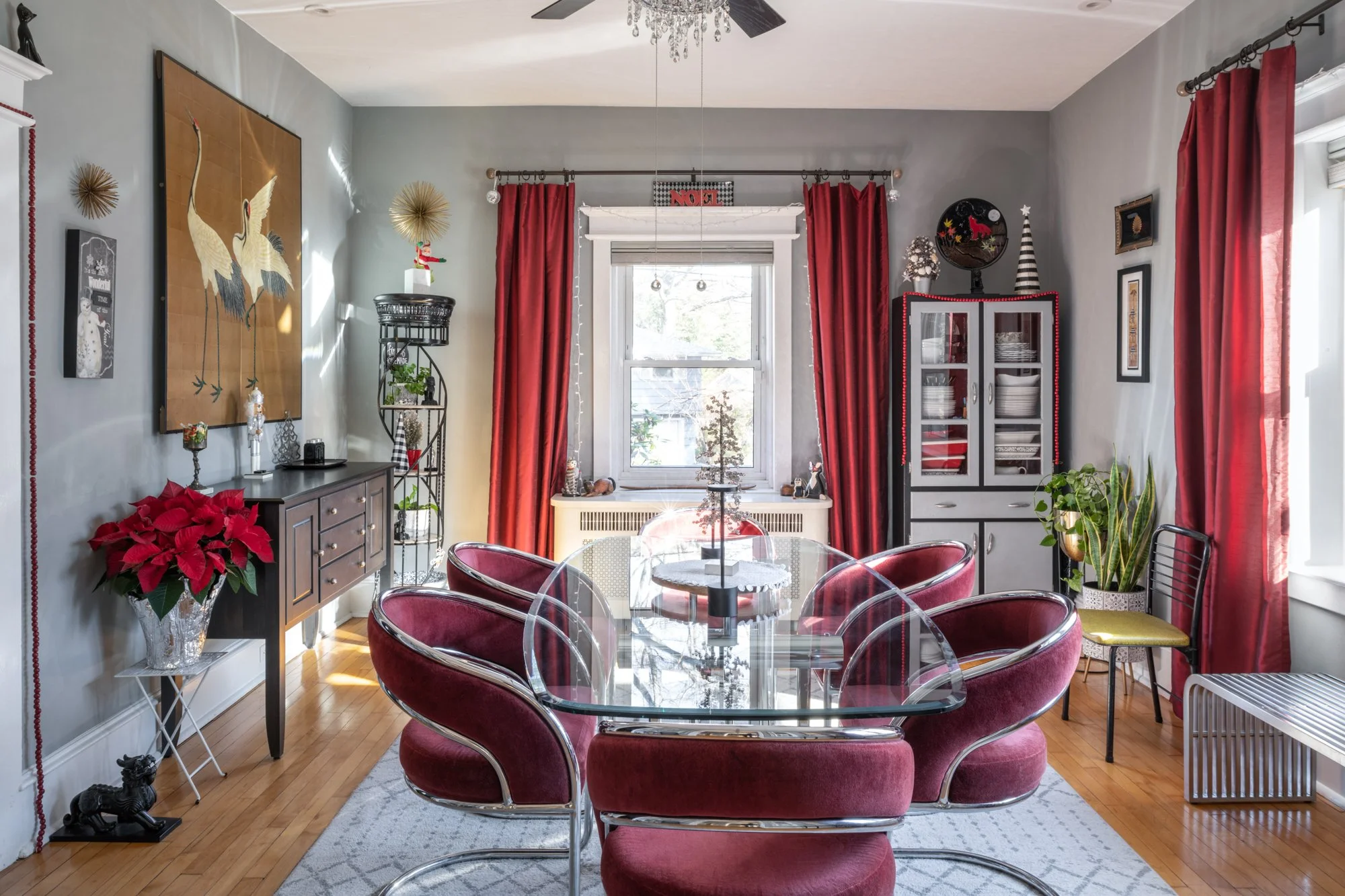 A dining room decorated for Christmas with red curtains, a poinsettia plant, and festive decorations, including a display on top of a cabinet and a chandelier hanging from the ceiling.