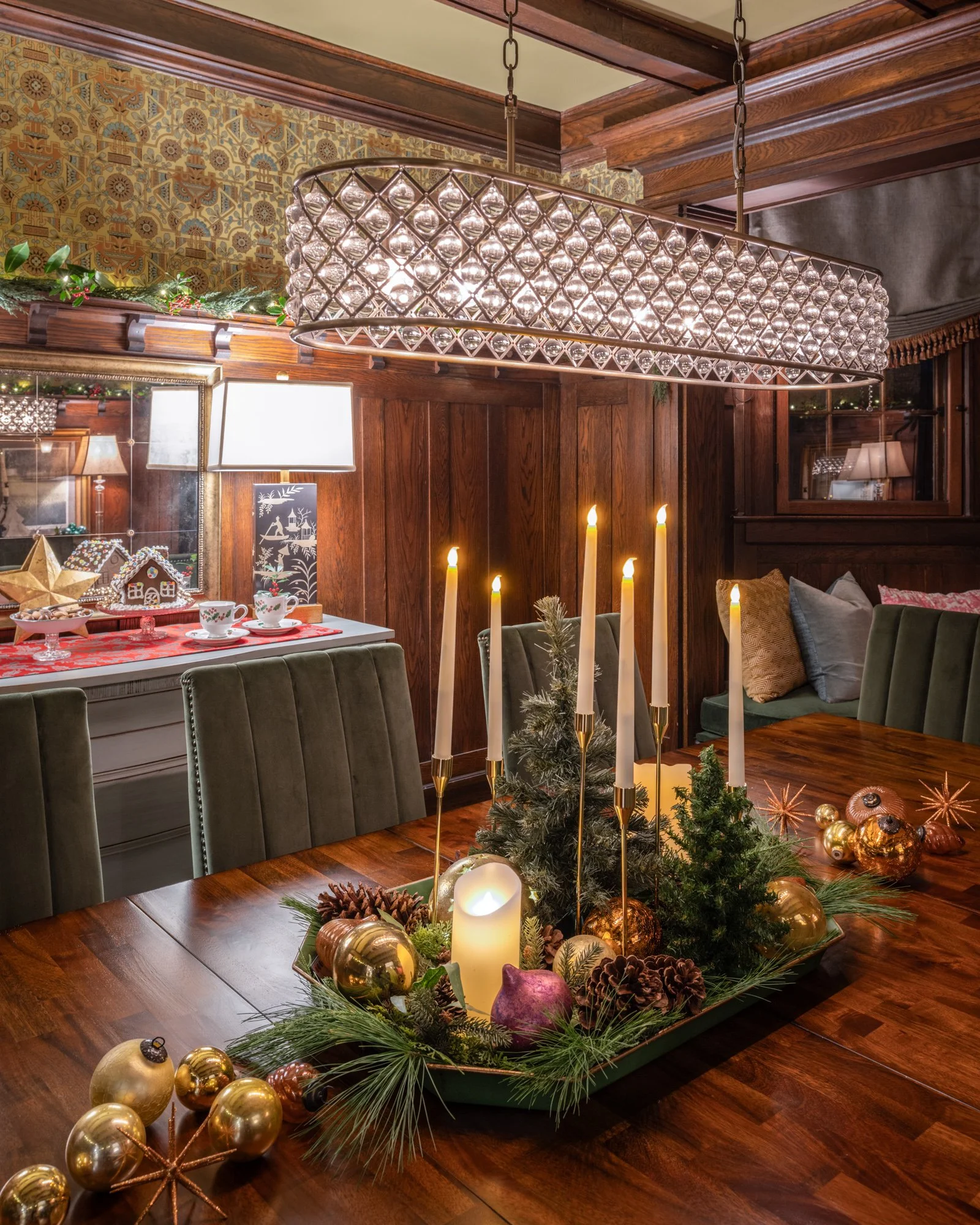 A warm holiday scene in a dining room featuring a wooden table decorated with a centerpiece of pine branches, candles, gold and purple ornaments, and pinecones. In the background, there are decorative candles, plates, cups, and holiday decor, with wa