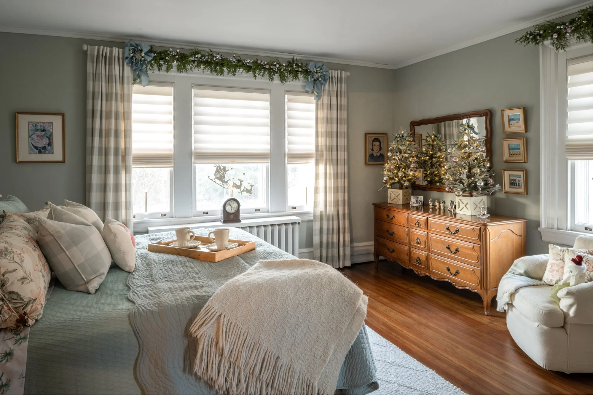 Living room decorated for Christmas with a Christmas tree on a wooden dresser, holiday garland with blue bows and ornaments above the windows, framed pictures on the wall, curtains, and a pastel green wall.
