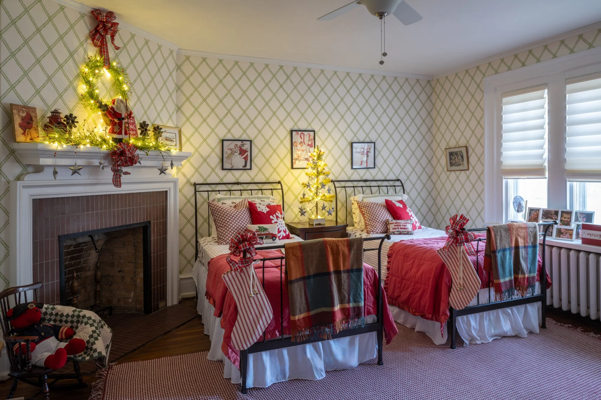 A cozy bedroom decorated for Christmas with three beds covered in red, plaid, and white bedding. Each bed has festive pillows and stockings hanging from the headboards. A small decorated Christmas tree with lights and stars stands on the nightstand b