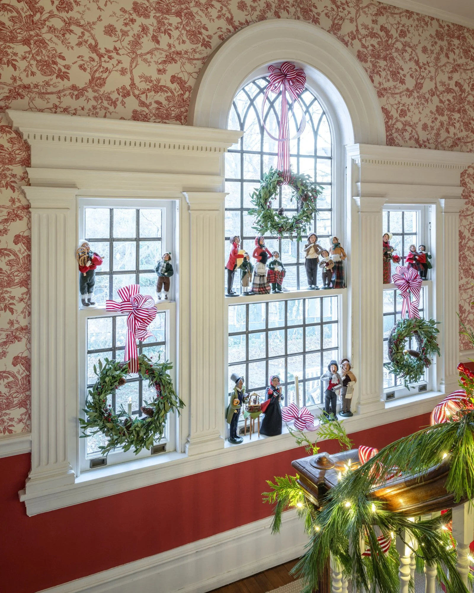 Decorative Christmas display with figurines of people, wreaths, ribbons, and greenery arranged on window sills and around a large window in a room with floral wallpaper and white trim.