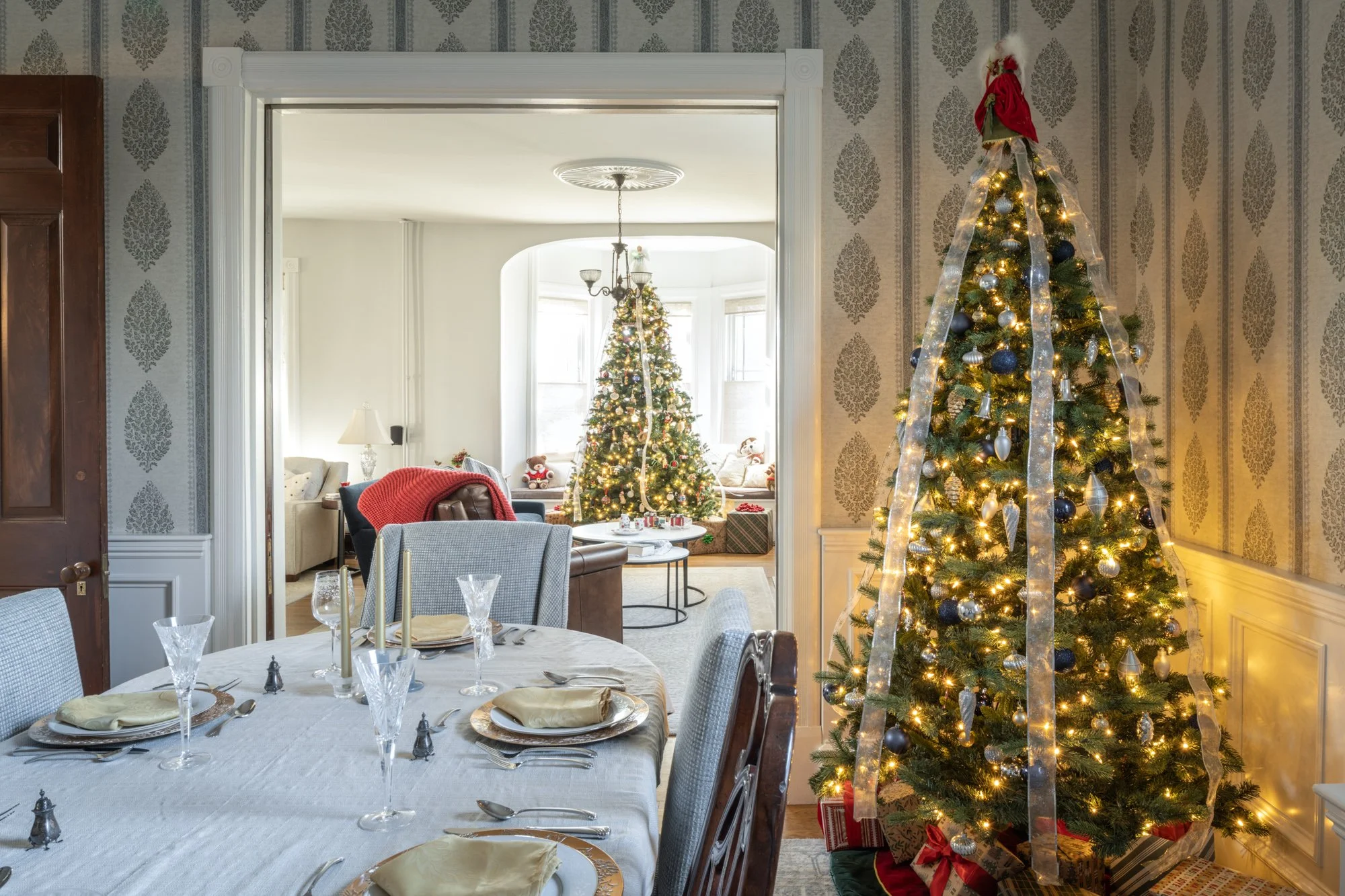 A decorated Christmas tree with ornaments, ribbons, and lights in a living room next to a dining table set for a meal.