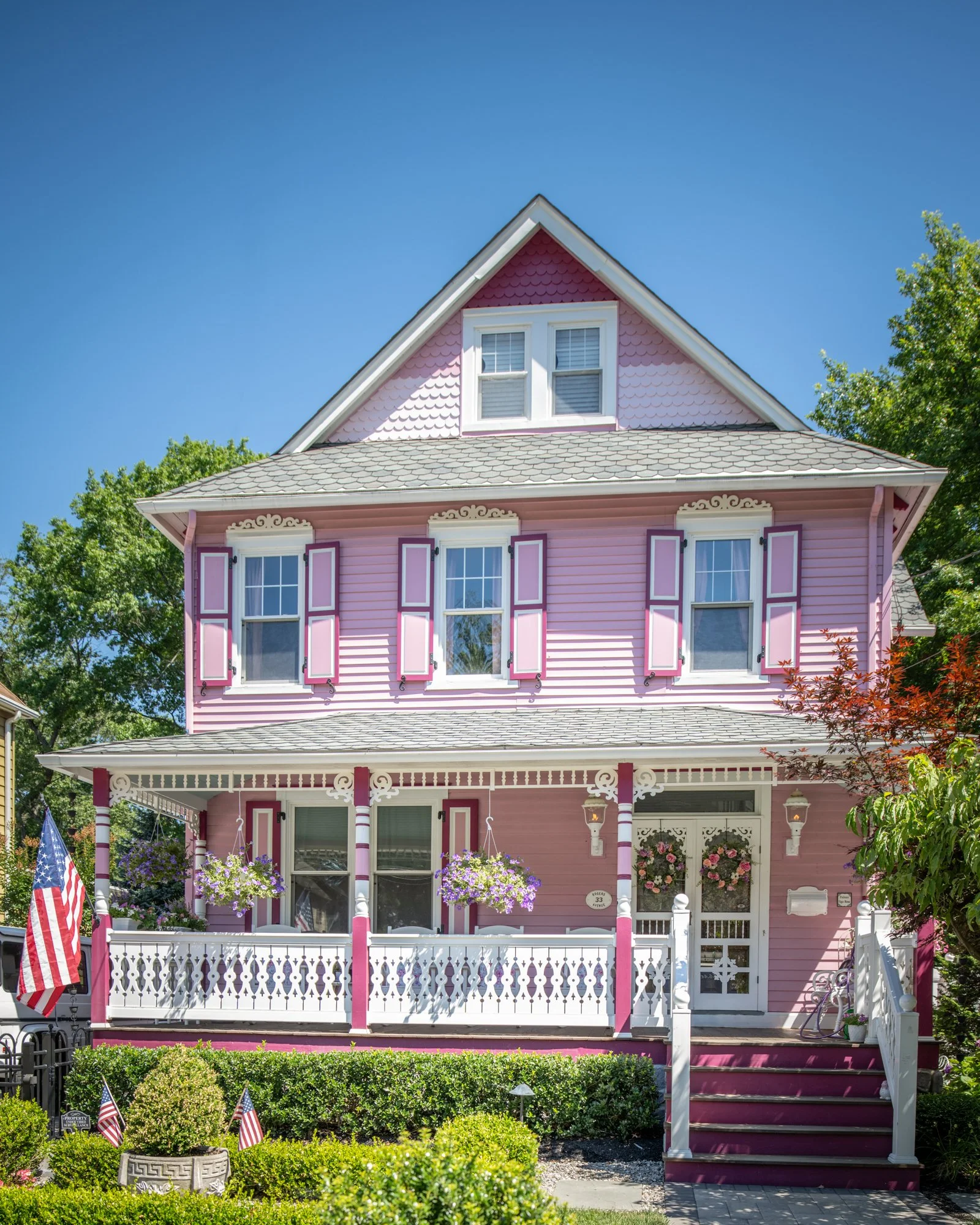A pink Victorian house with a front porch decorated with hanging flower baskets, American flags, and pink shutters. The house has multiple windows and a gable roof with shingles, and is surrounded by greenery and trees under a clear blue sky.