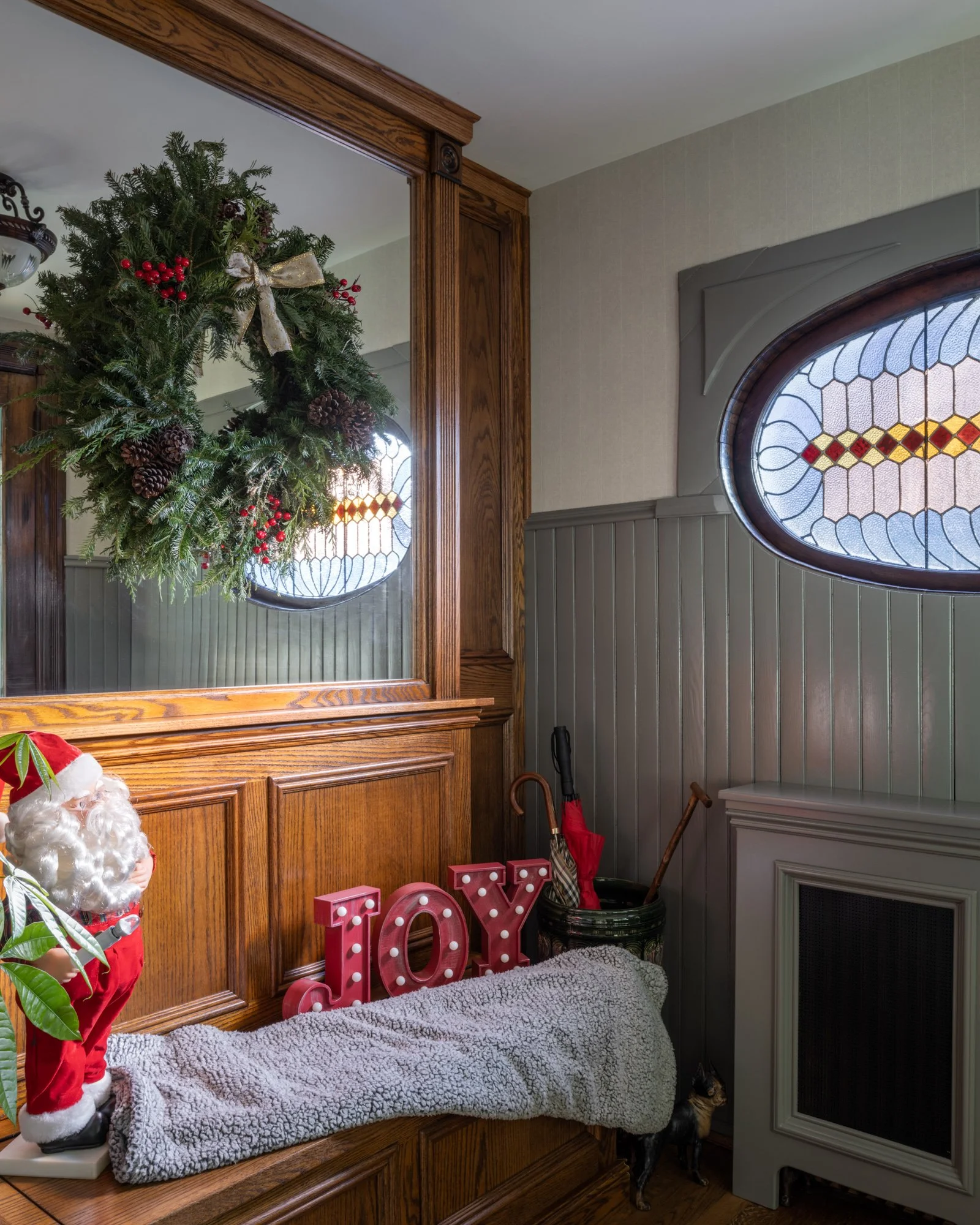 Indoor Christmas scene with a large mirror, a decorated Christmas wreath, a Santa Claus figure, pink 'LOVE' marquee letters, and a cat sitting on a gray textured blanket on a wooden bench.