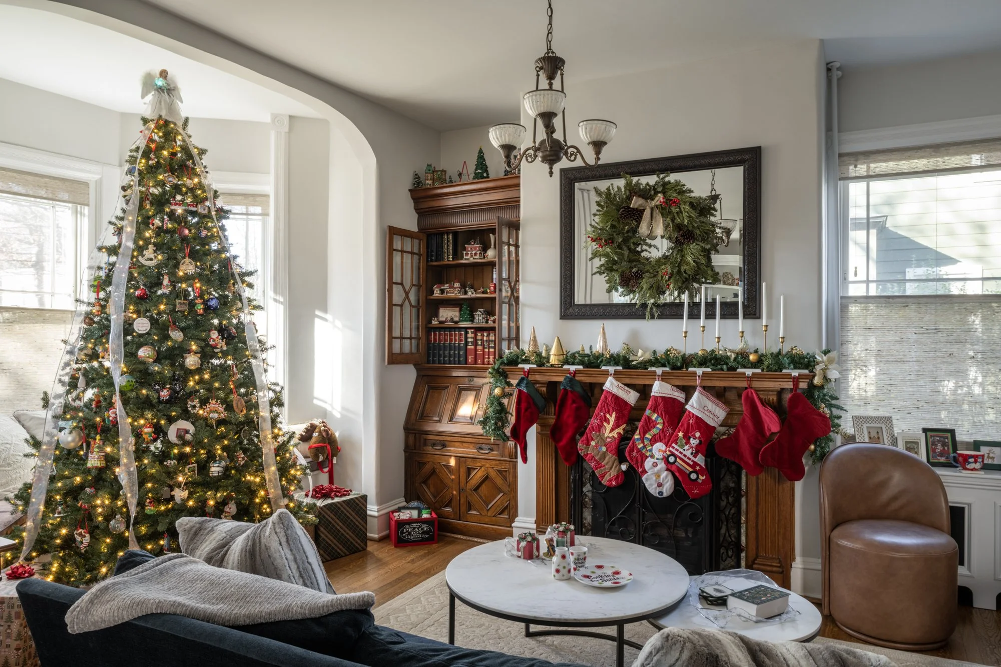 Living room decorated for Christmas with a lit Christmas tree, stockings hanging on a fireplace, wreath on the mirror, and holiday decorations on the mantel and table.
