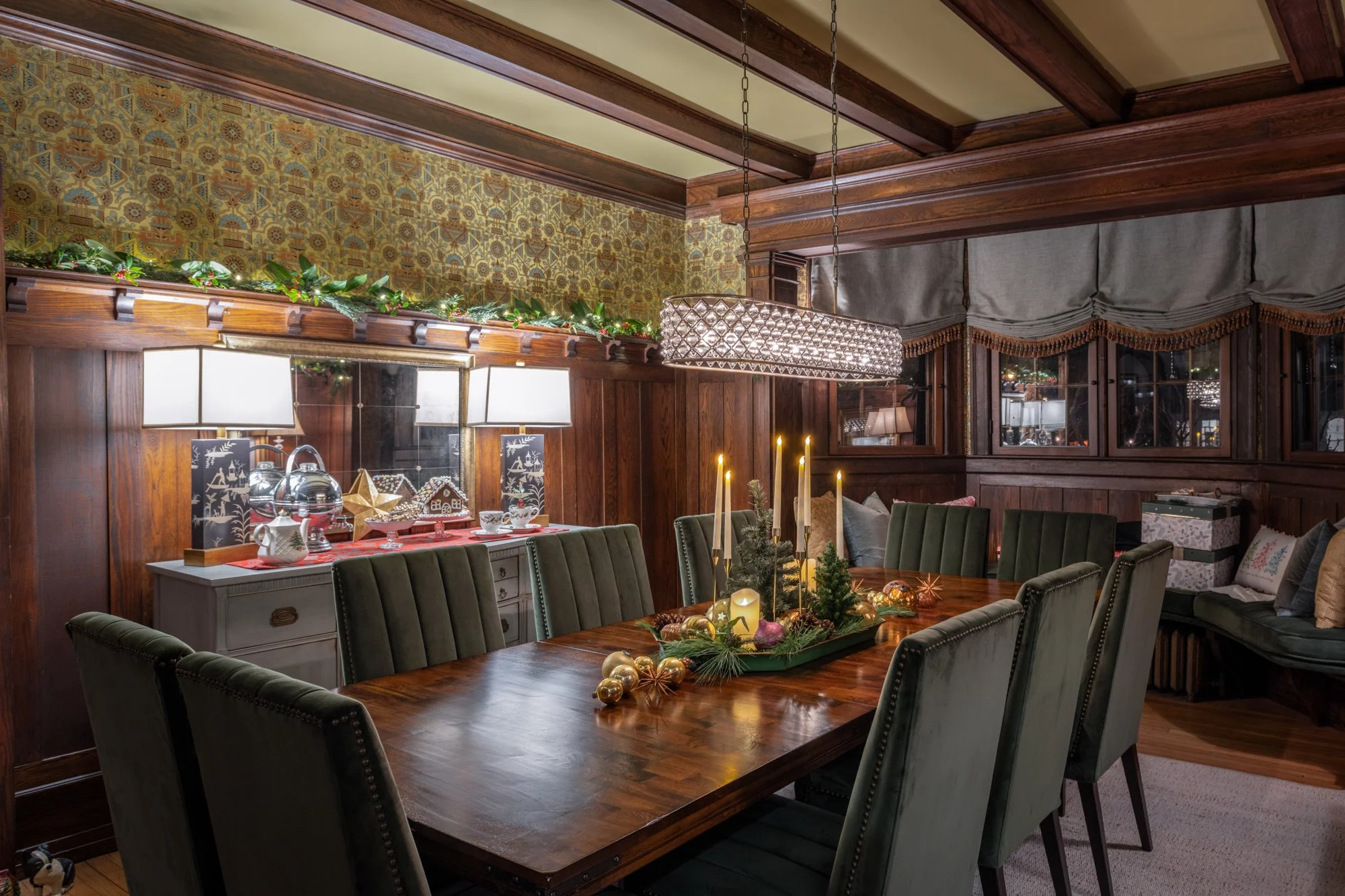 A cozy dining room decorated for Christmas with a wooden table, green chairs, a festive centerpiece, lit candles, and holiday decorations. The room has dark wood paneling, a patterned wallpaper, and a chandelier hanging from the ceiling.