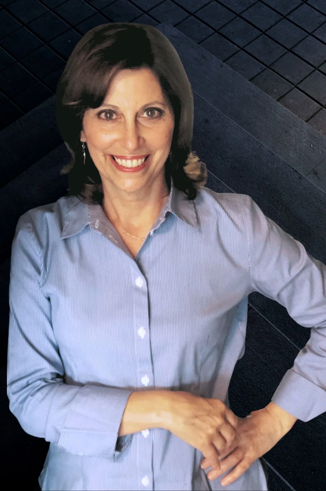 A woman with dark brown hair, wearing a light blue striped shirt, smiling and standing with her hands on her hips against a dark blue wooden floor background.