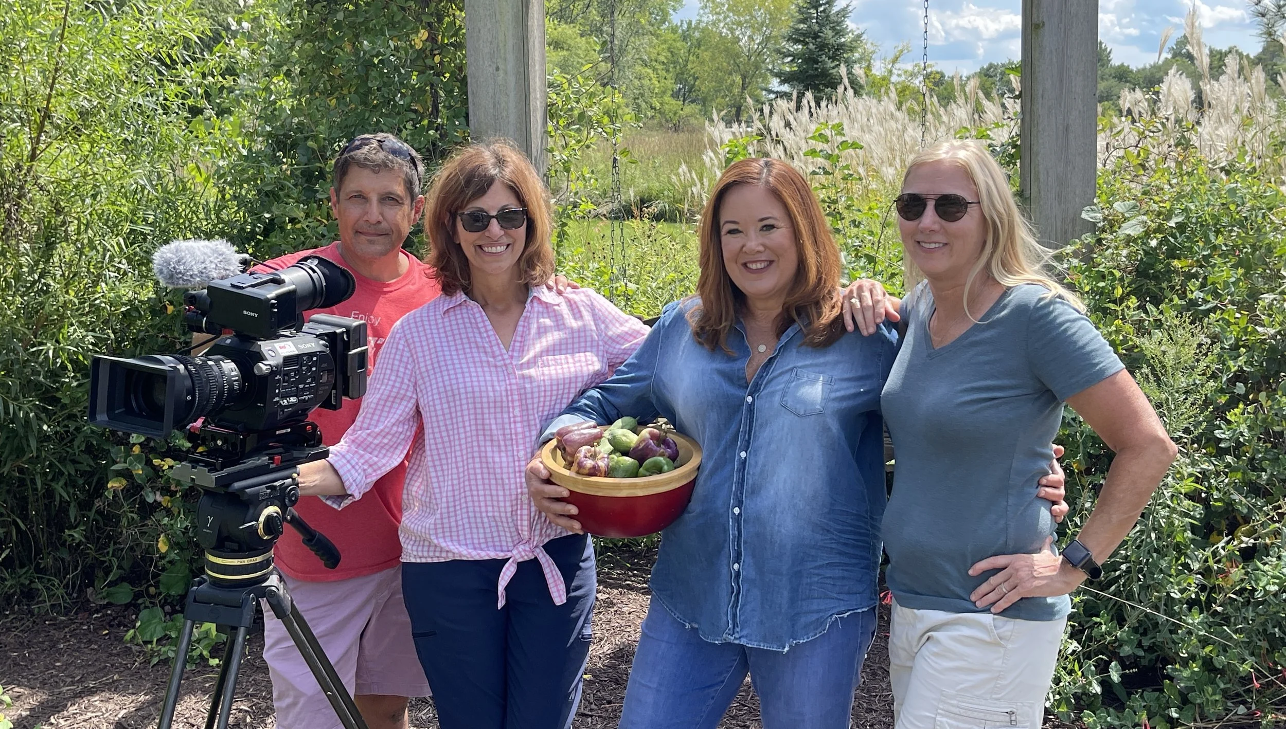 Four women and one man outdoors with a camera on a tripod, surrounded by green bushes and trees, one woman holding a bowl of fresh figs.