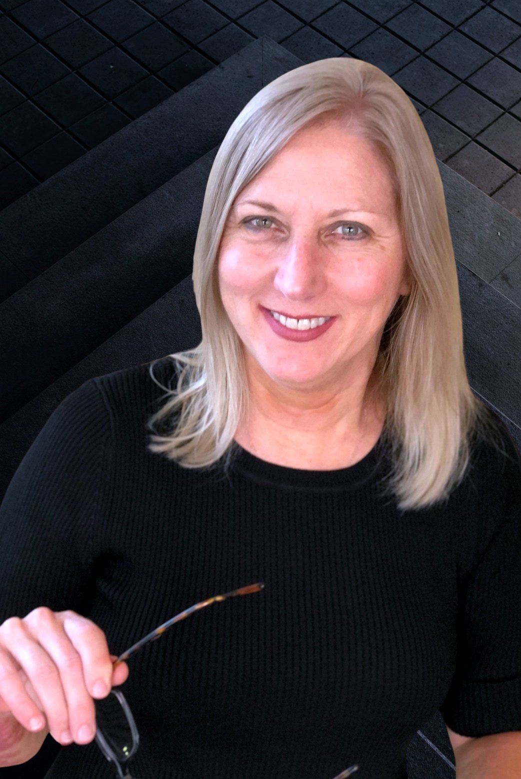 Smiling woman with blonde hair wearing a black top, holding eyeglasses, sitting on a dark tiled surface.