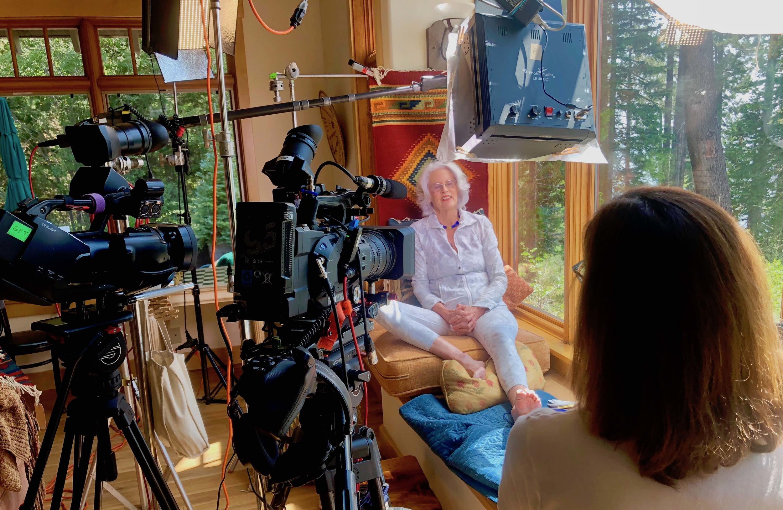 An elderly woman sitting on a window seat being filmed by a camera crew for an interview or video shoot, with large windows showing a wooded outdoor scene.