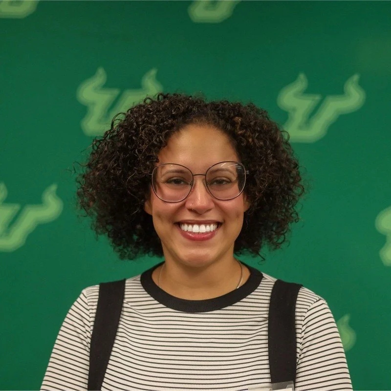A woman with curly hair and glasses smiling, standing against a green background with abstract plant-like designs.